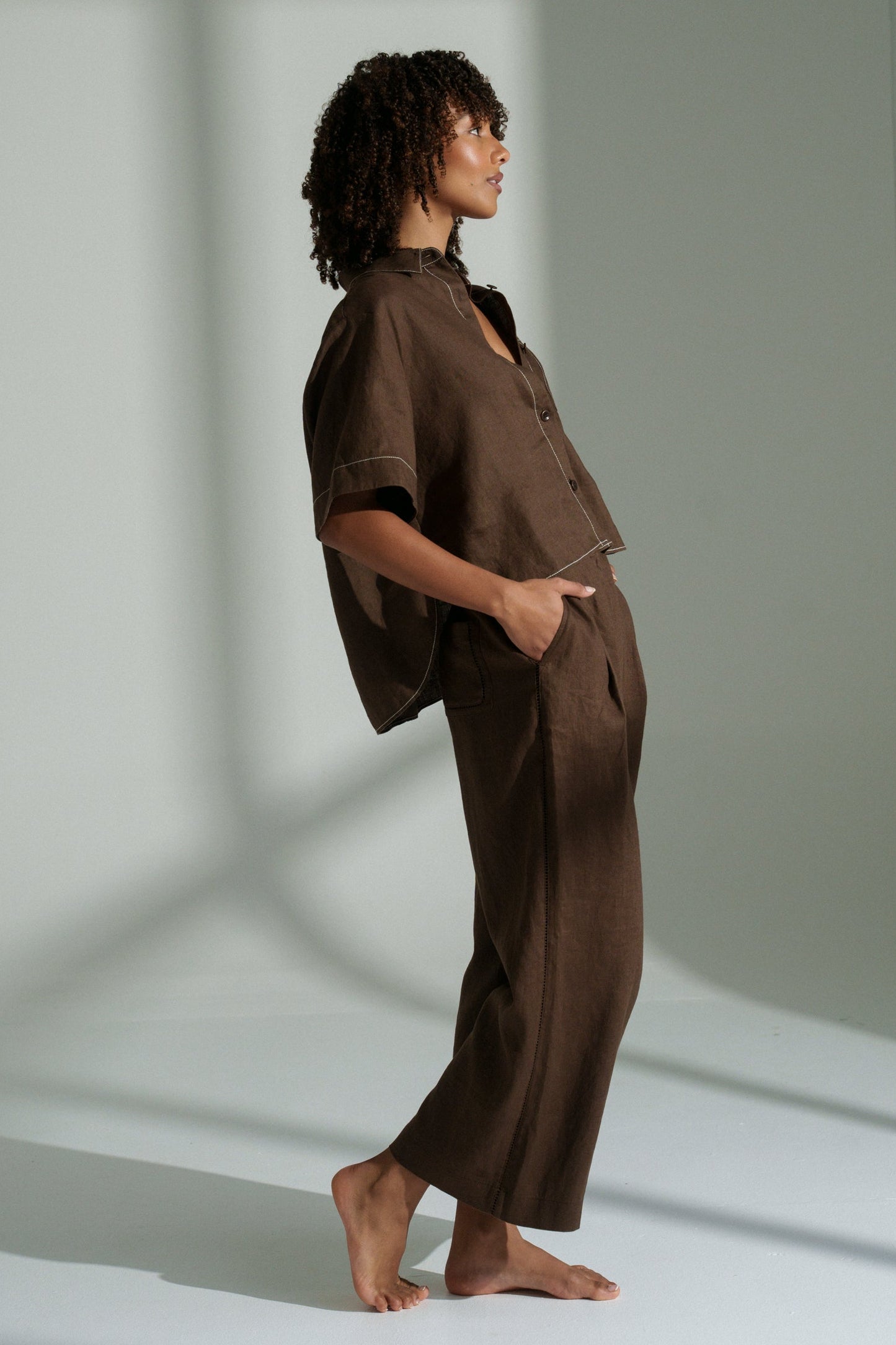 LILLY PILLY woman wearing Rika linen pants and Nika linen shirt in chocolate brown standing in a softly lit room with shadows on the wall.