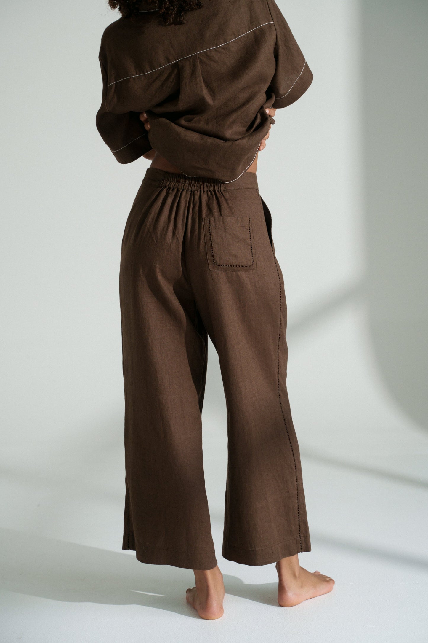 LILLY PILLY woman wearing Rika linen pants in chocolate brown with on a white background, back view.