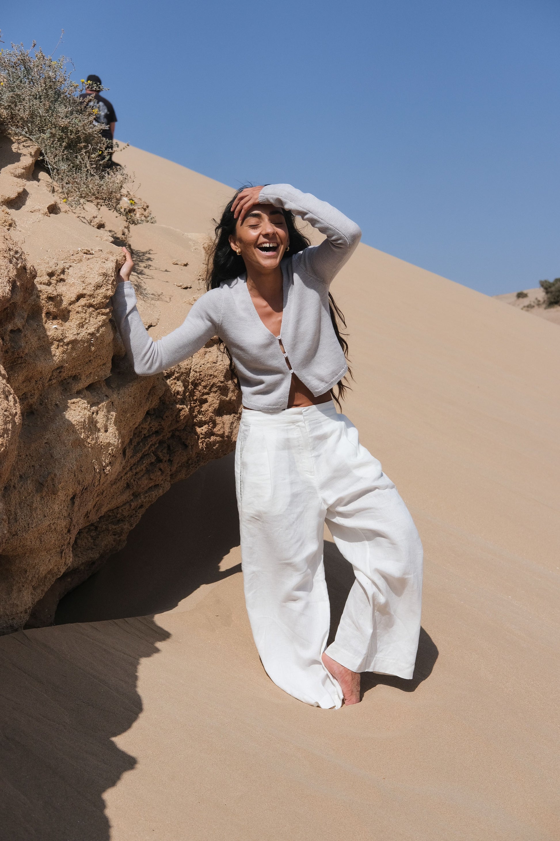 LILLY PILLY woman in ivory Rika linen pants standing on sand dunes with a clear blue sky.