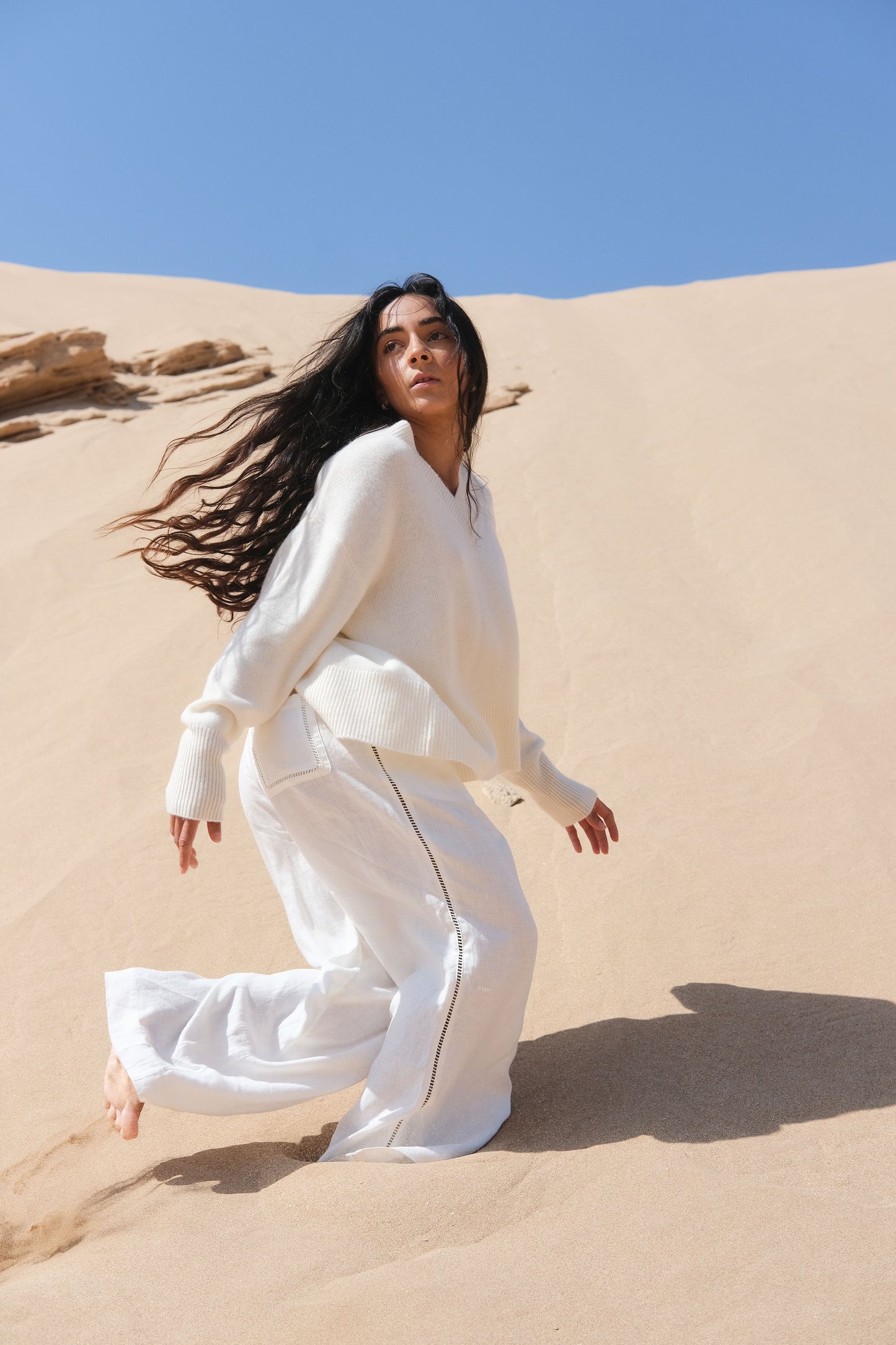 LILLY PILLY woman in ivory Rika linen pants standing on sand dunes with a clear blue sky.