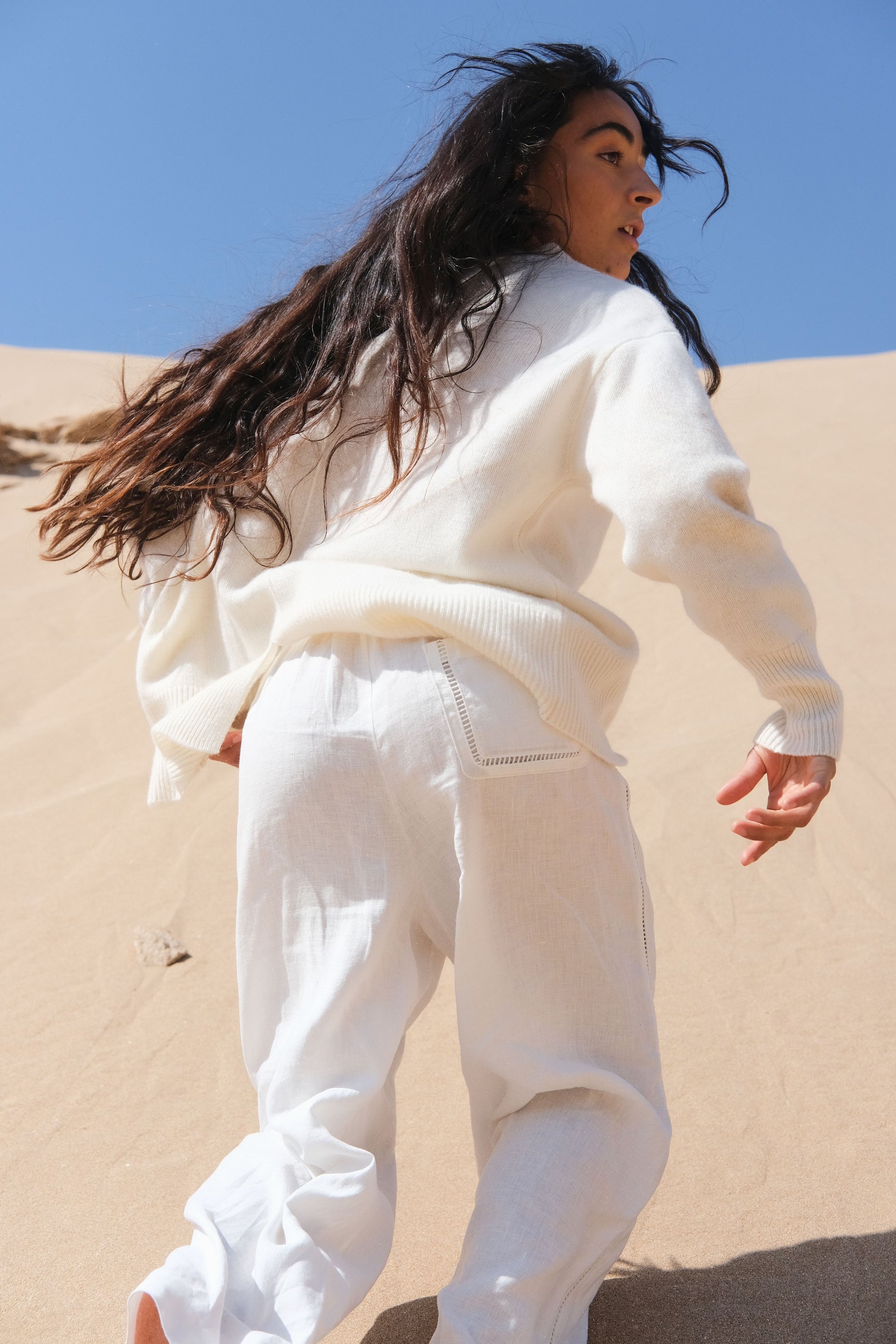 LILLY PILLY woman in ivory Rika linen pants standing on sand with a blue sky background