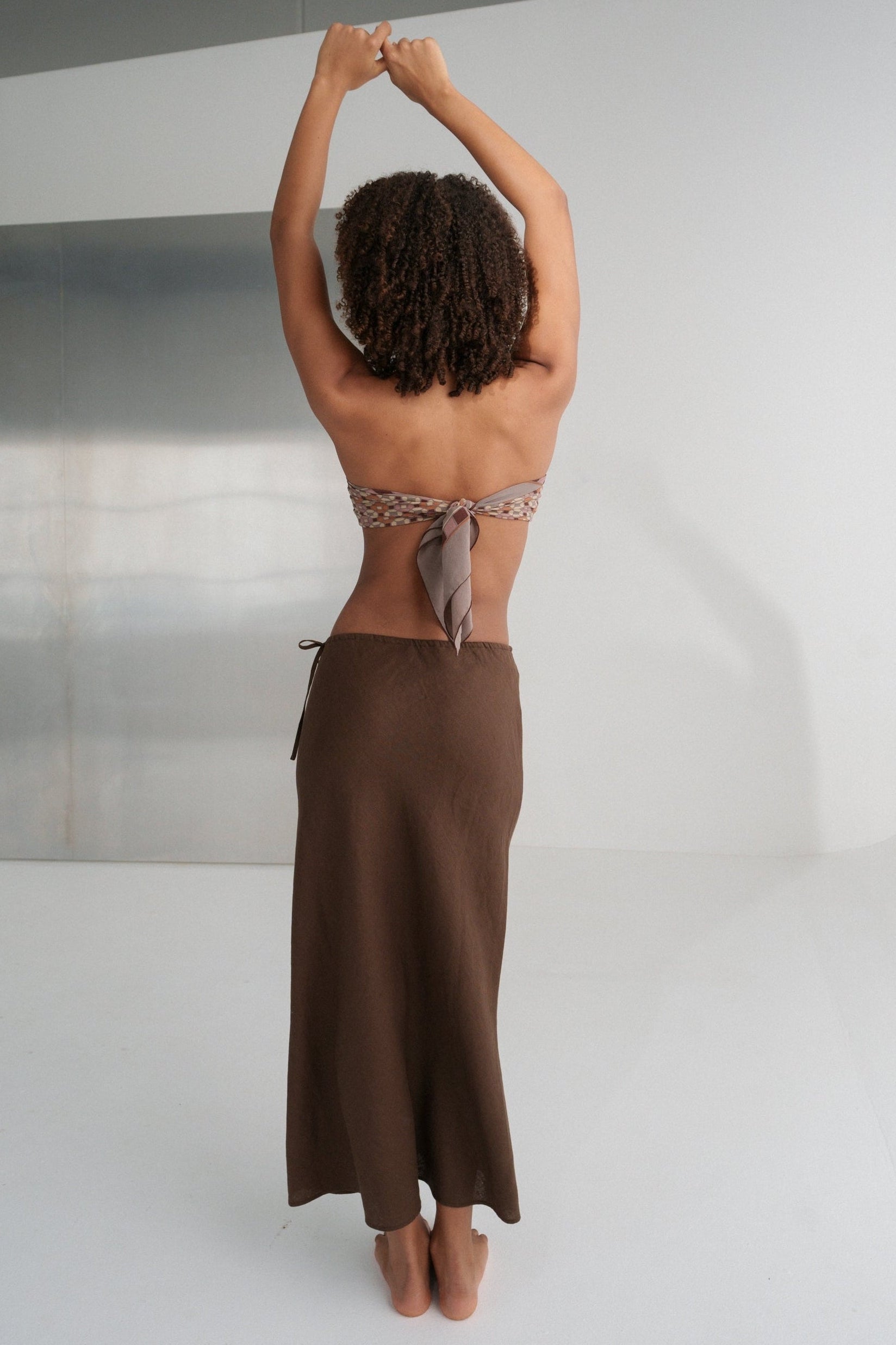 LILLY PILLY woman wearing Riley bias cut linen skirt  in chocolate brown standing with arms raised against a white wall.