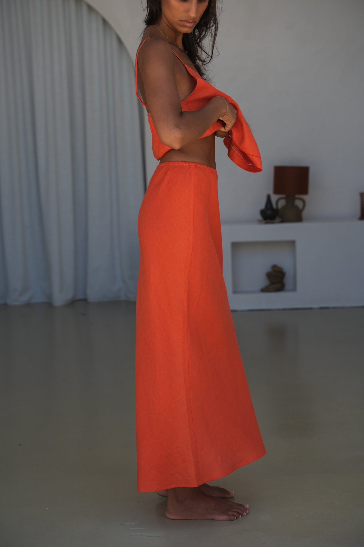 LILLY PILLY woman wearing Riley linen skirt in Spritz standing in a minimalistic room with white walls and a curtain.