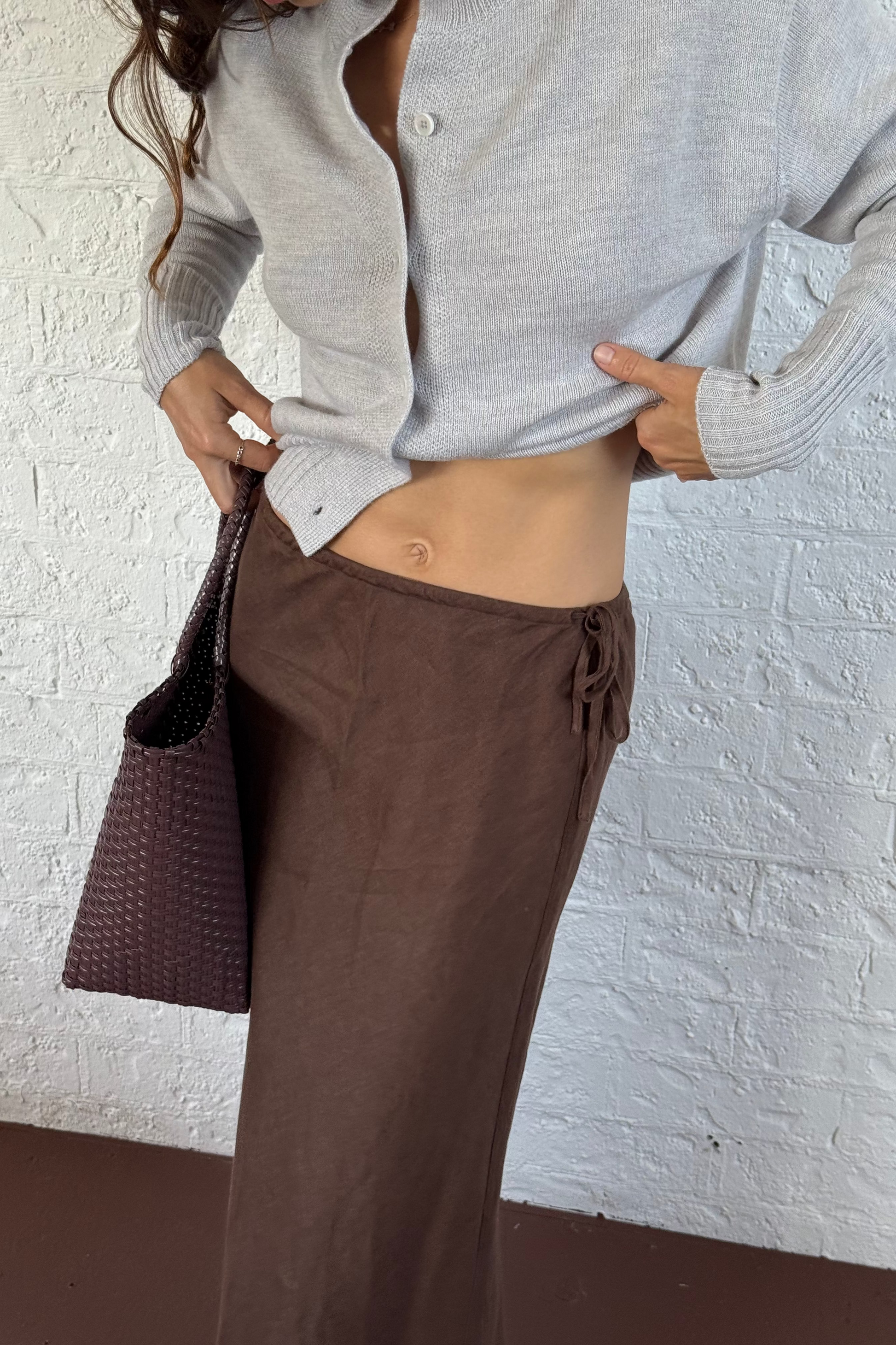 LILLY PILLY werson wearing a fog gray button-up Taylor merino knit cardigan top and chocolate brown Riley linen skirt, holding a woven bag against a white brick wall.