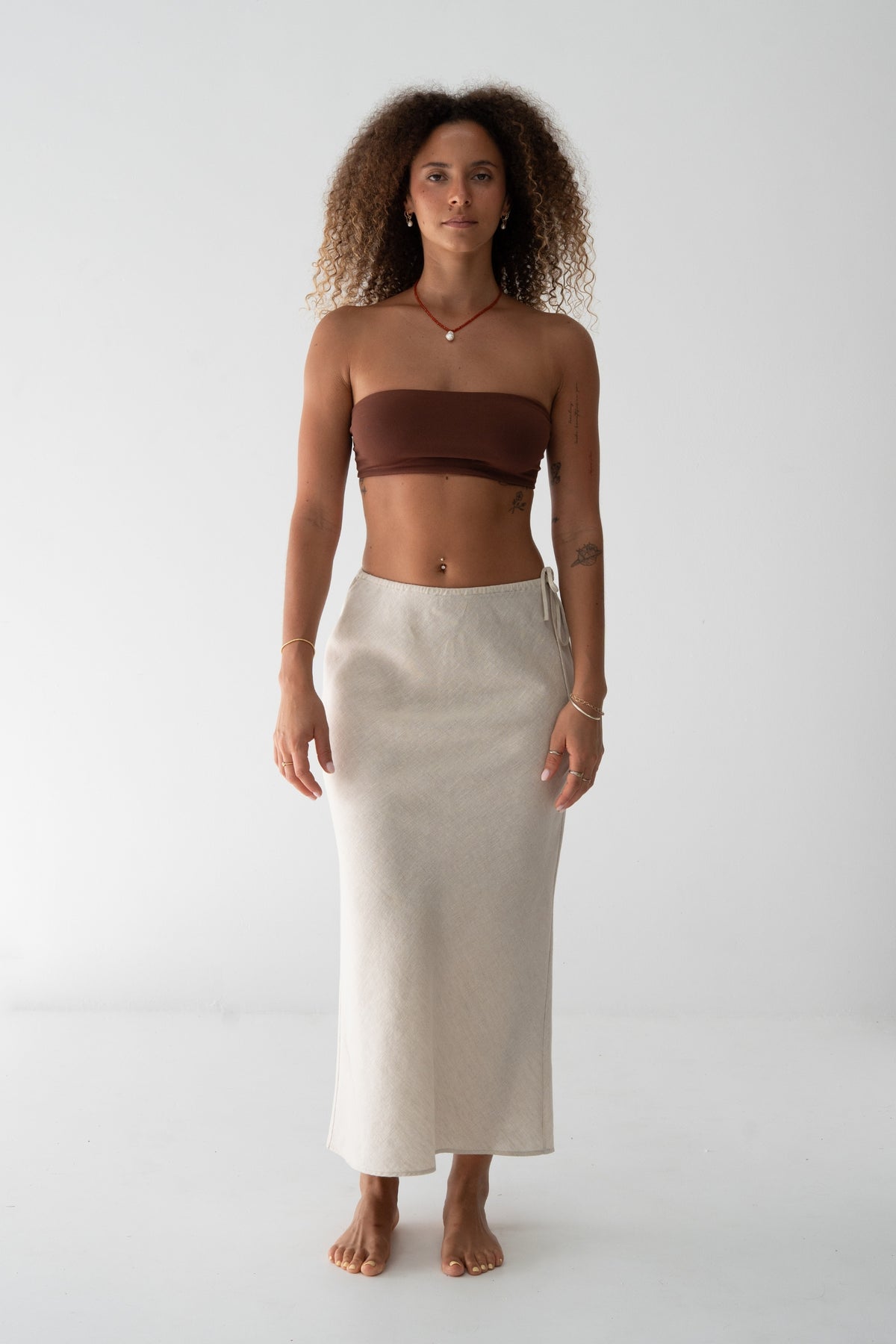 Riley Bias Linen Midi Skirt in Oatmeal - front view, bias-cut midi length, side tie detail, worn with chocolate bandeau top, gold jewellery - Lilly Pilly Collection