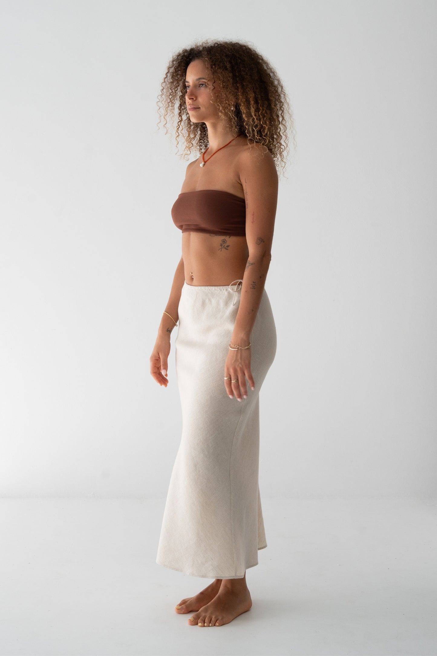 Riley Bias Linen Midi Skirt in Oatmeal - side view, bias-cut midi length, side tie detail, worn with chocolate bandeau top, gold jewellery - Lilly Pilly Collection