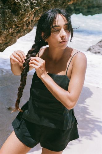 Rosa Linen Silk Cami in Black - woman holding braid, square neckline and spaghetti straps, styled with matching black linen silk shorts, rocky beach and ocean backdrop - Lilly Pilly Collection