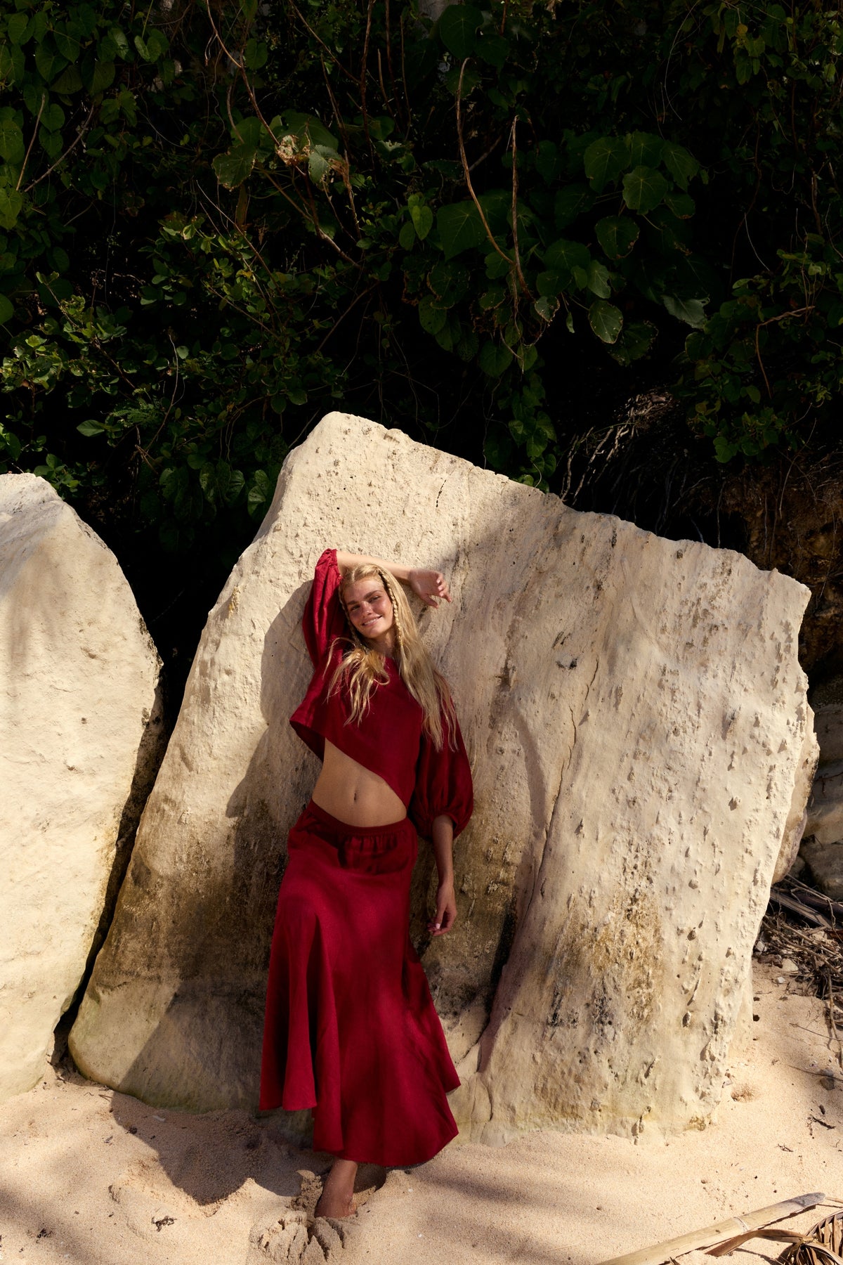 Stella Linen Maxi Skirt in Cherry Red - full length, woman leaning against rock wearing matching Cherry Red linen puff-sleeve crop top, coastal setting - Lilly Pilly Collection