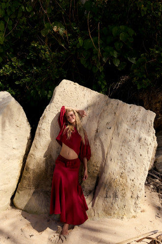 Stella Linen Maxi Skirt in Cherry Red - full length, woman leaning against rock wearing matching Cherry Red linen puff-sleeve crop top, coastal setting - Lilly Pilly Collection