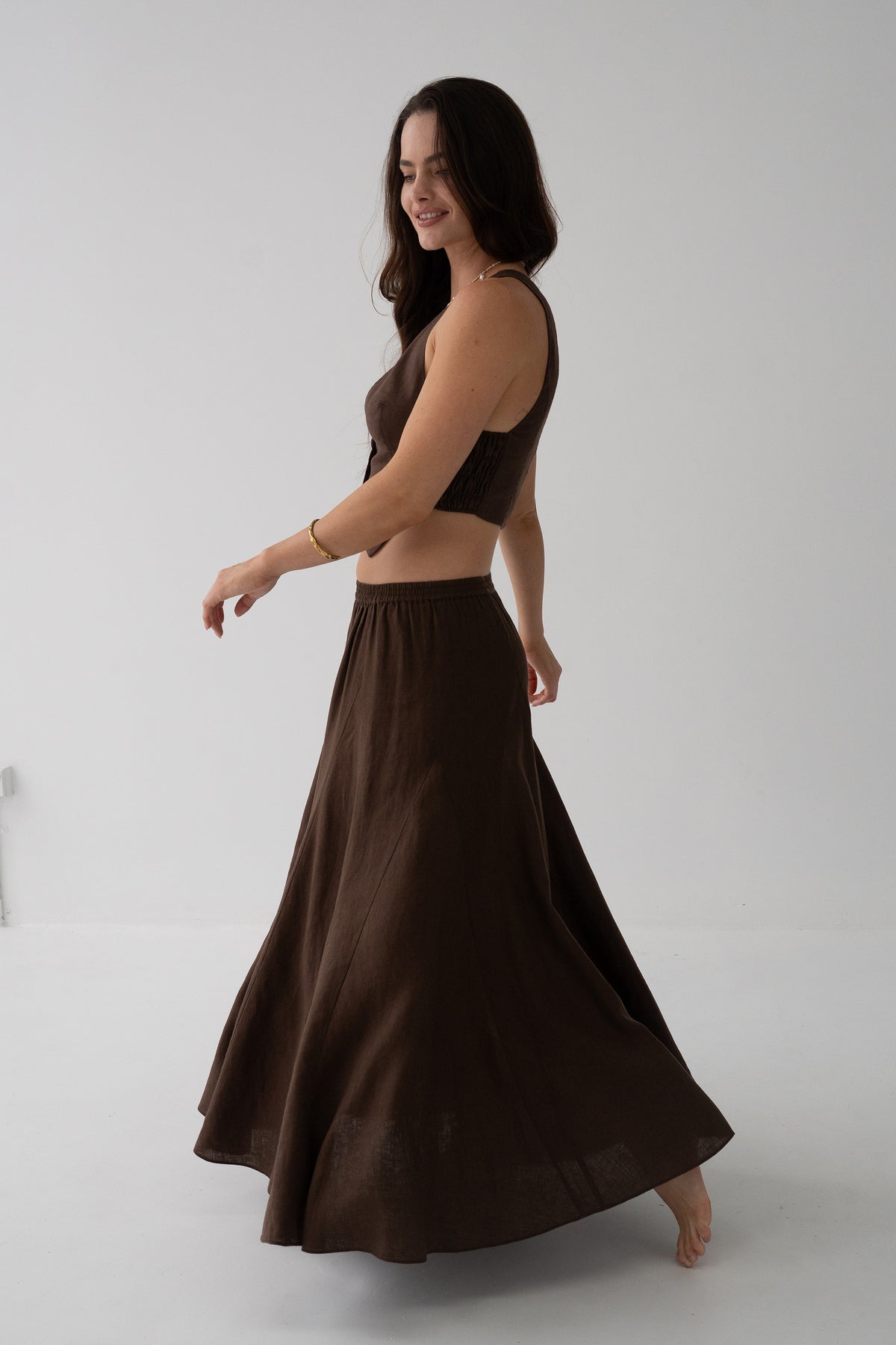 Stella Linen Maxi Skirt in Chocolate - side view, woman wearing Chocolate linen skirt with matching linen crop top, full A-line silhouette in motion, studio backdrop - Lilly Pilly Collection
