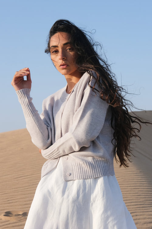 Woman in a light gray sweater and white pants standing on sand dunes with a clear blue sky.