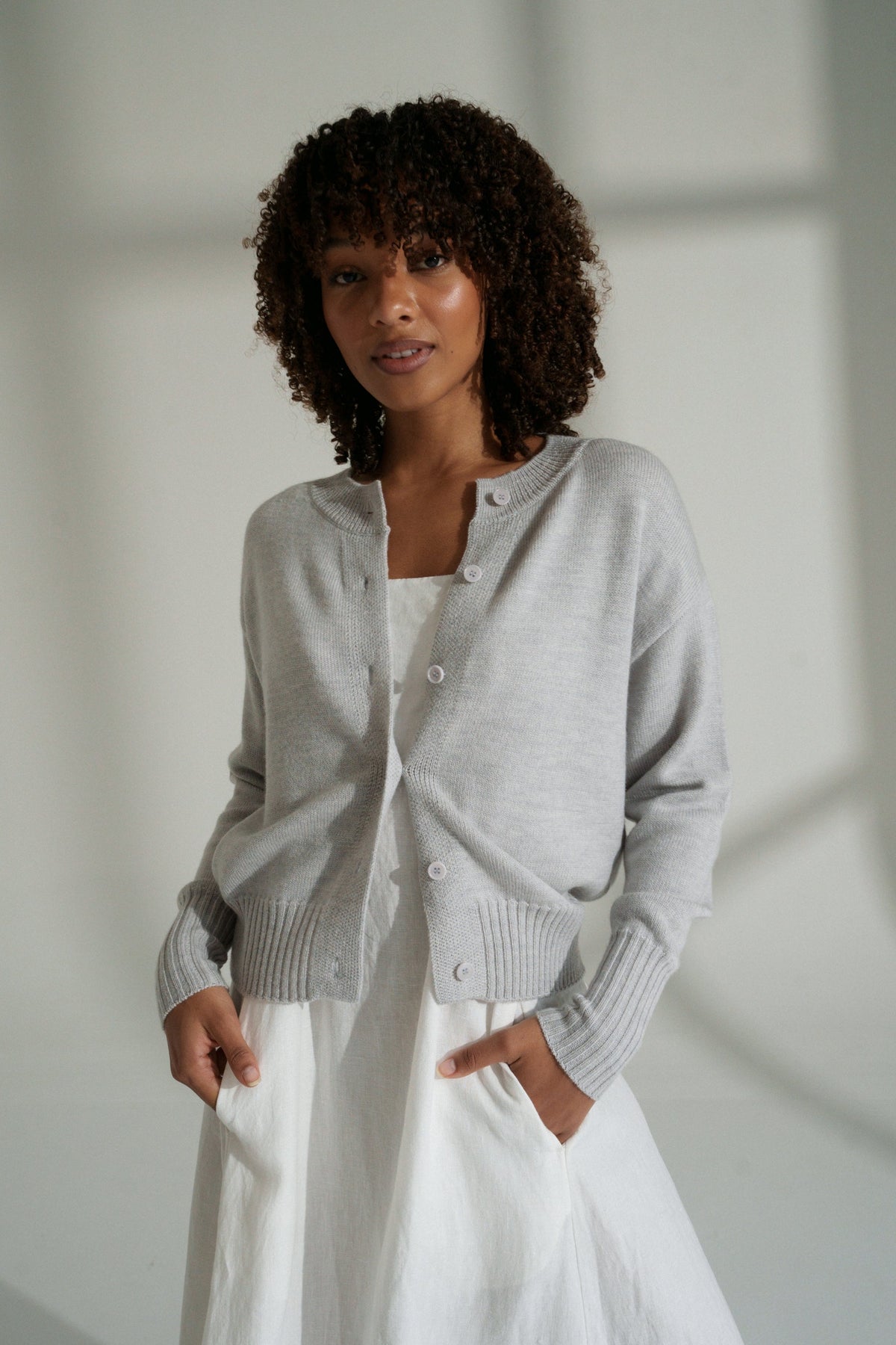LILLY PILLY woman wearing fog  coloured Taylor merino knit cardigan over a white dress against a neutral background
