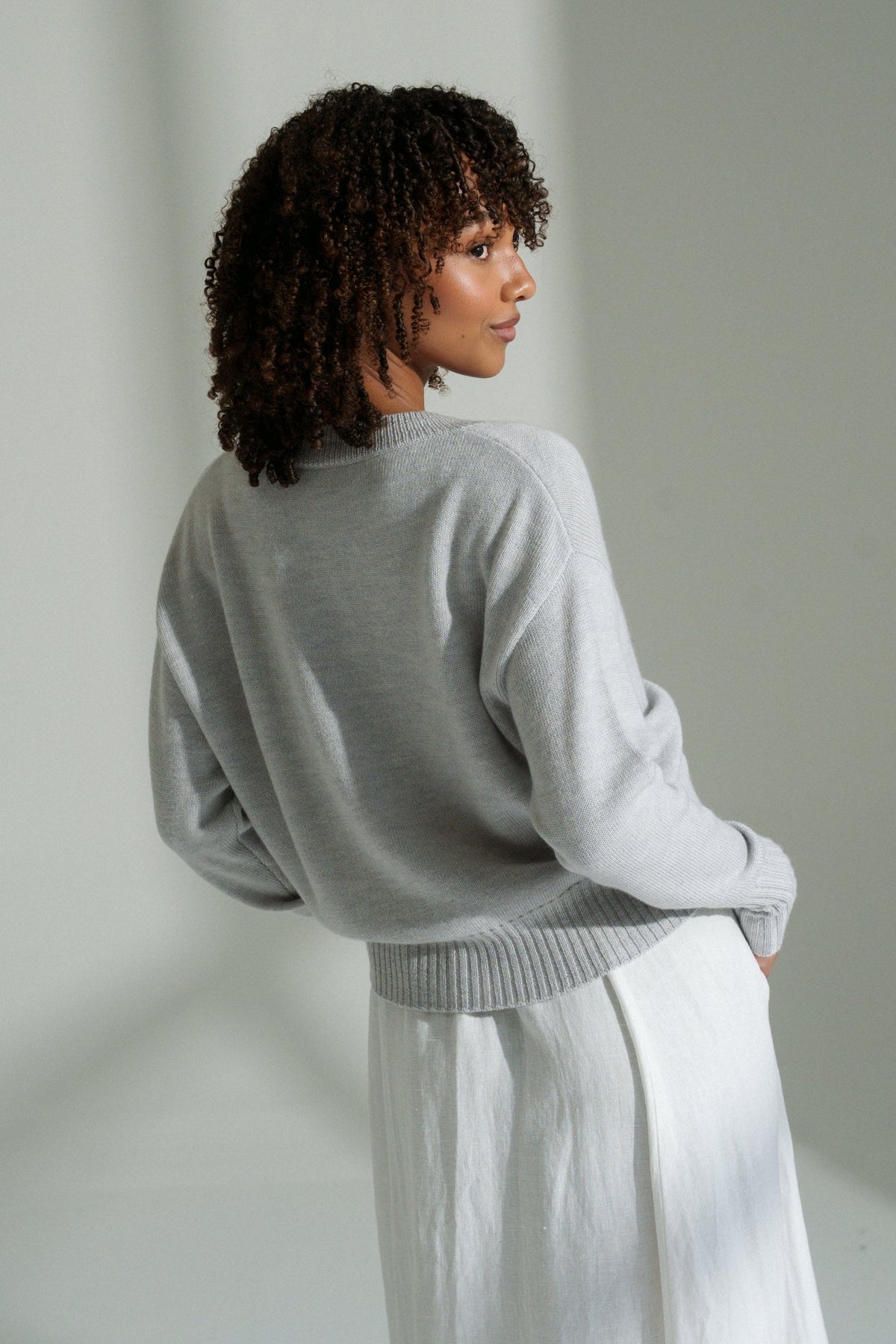 Back view of LILLY PILLY woman wearing fog coloured Taylor merino knit cardigan standing against a plain wall with soft shadows.