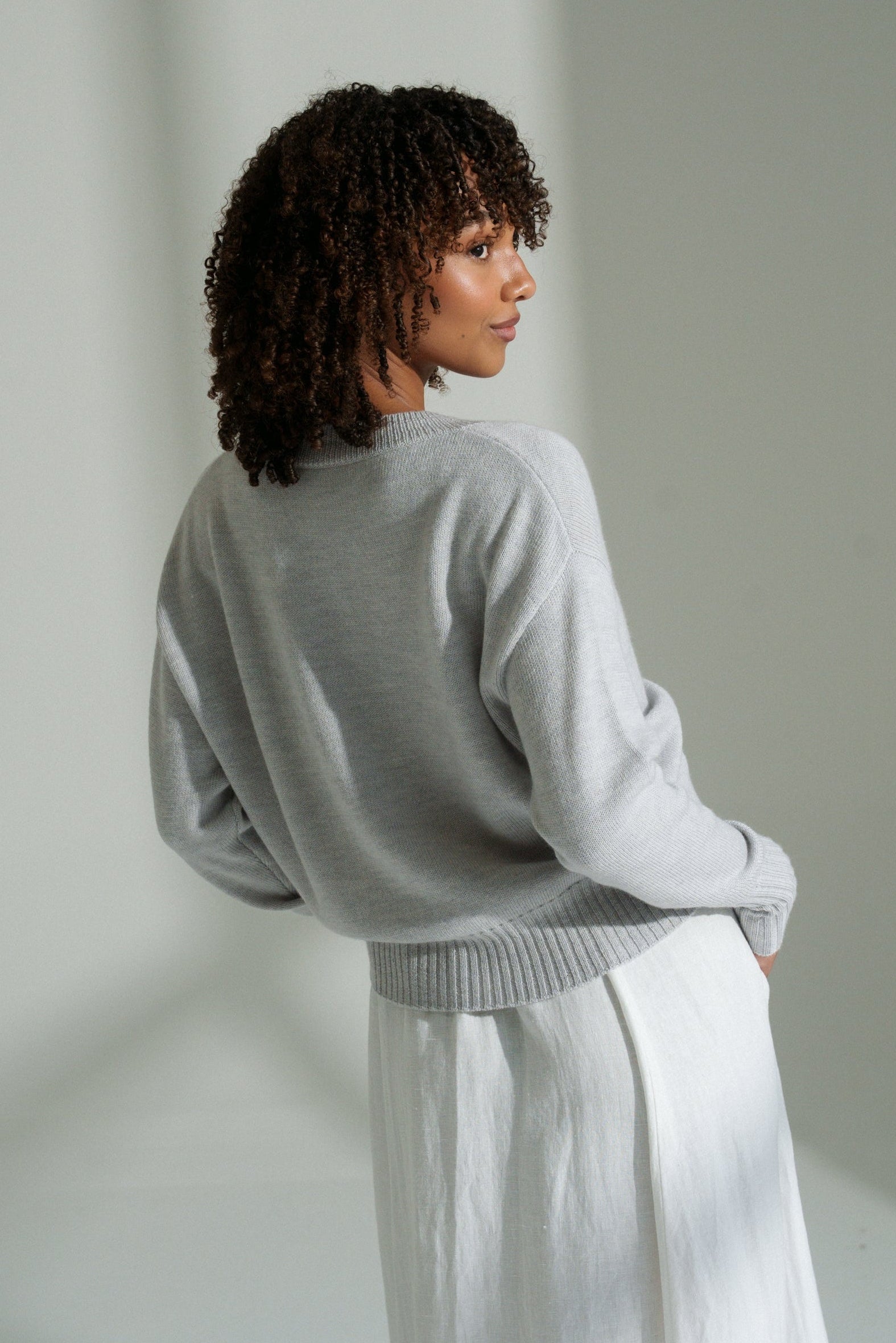 Back view of LILLY PILLY woman wearing fog coloured Taylor merino knit cardigan standing against a plain wall with soft shadows.