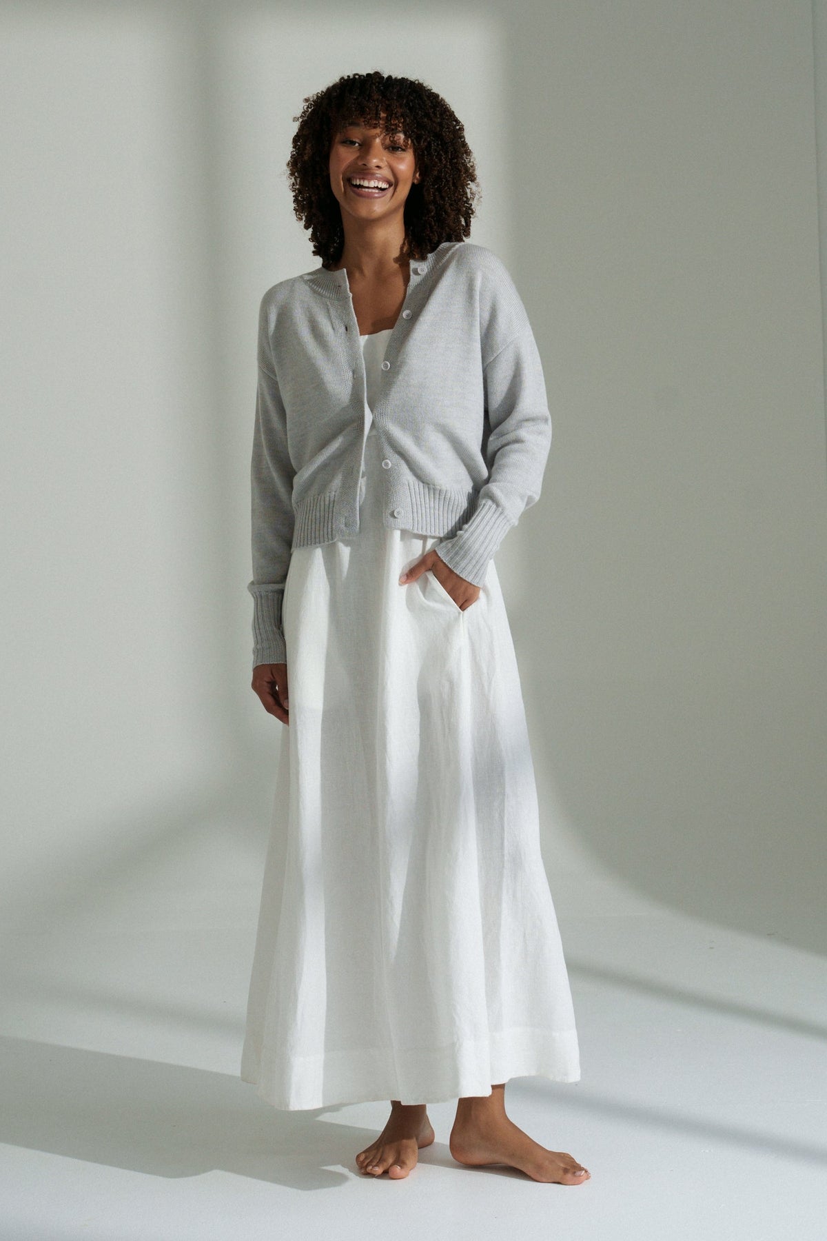 LILLY PILLY woman wearing fog coloured Taylor merino knit cardigan standing in a softly lit room.