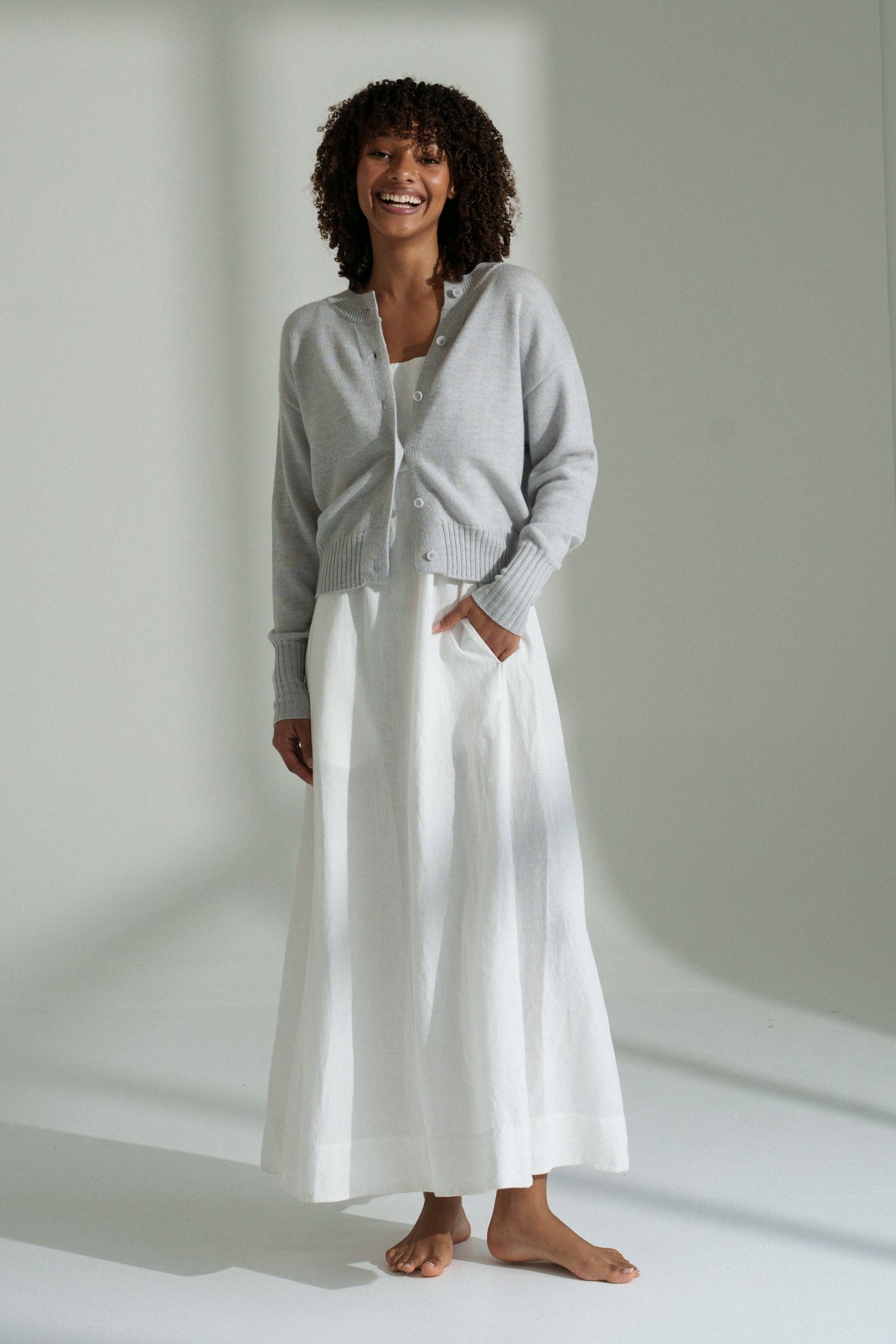 LILLY PILLY woman wearing fog coloured Taylor merino knit cardigan standing in a softly lit room.