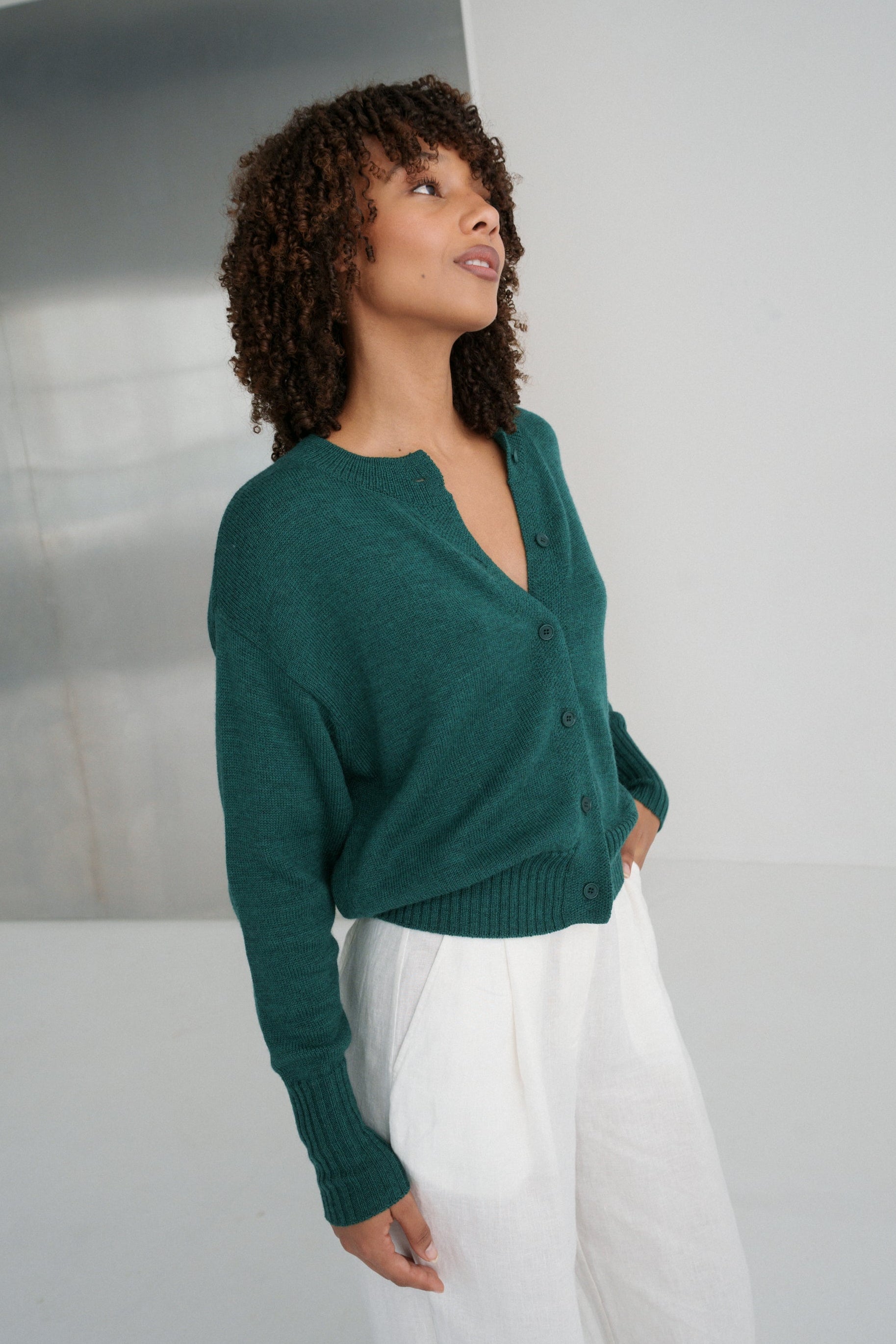 Side view of LILLY PILLY woman wearing pine green coloured Taylor merino knit cardigan against a plain background