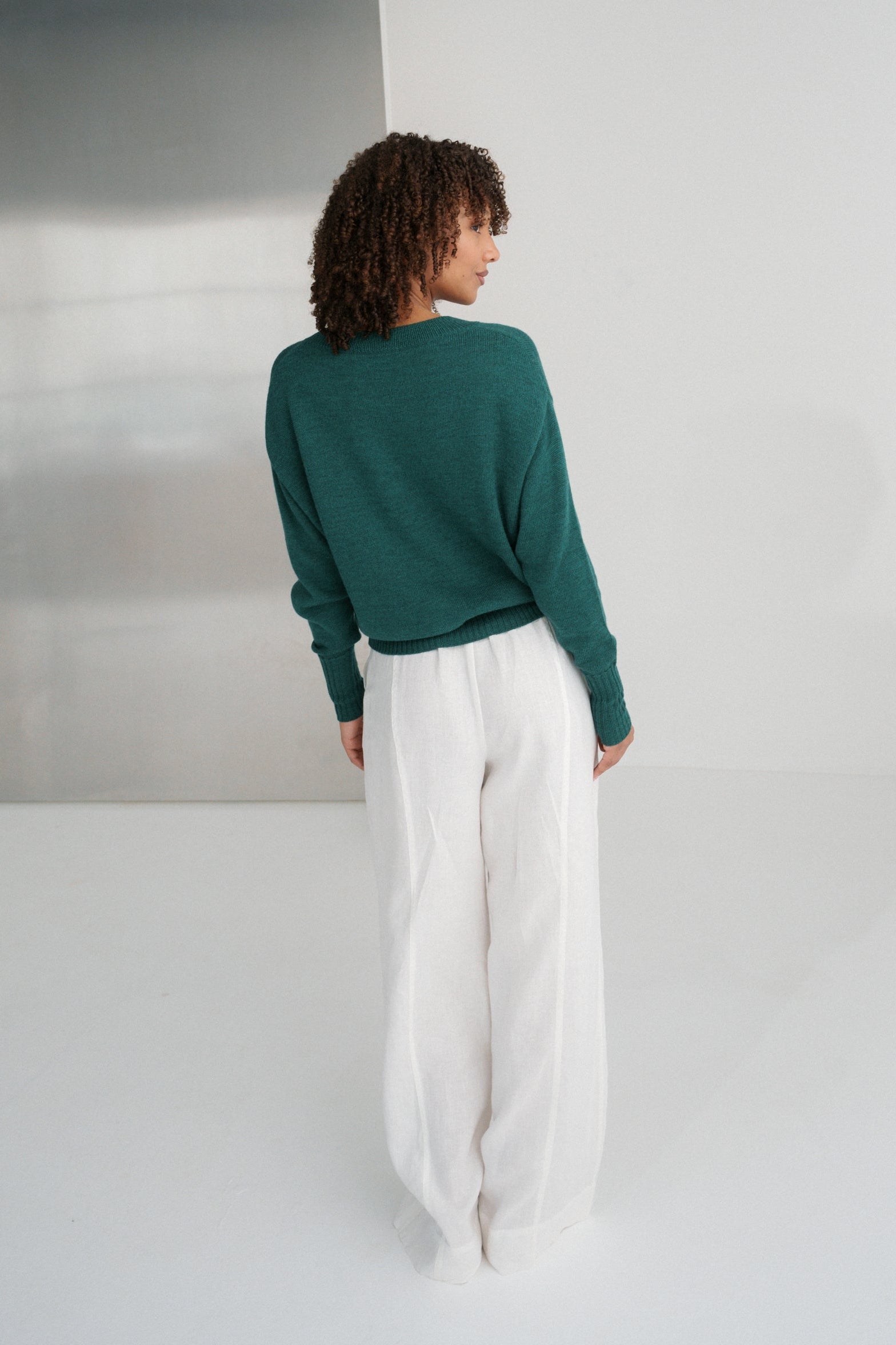 Back view of LILLY PILLY woman wearing pine green coloured Taylor merino knit cardigan and white pants standing in a minimalistic room with a metallic wall.