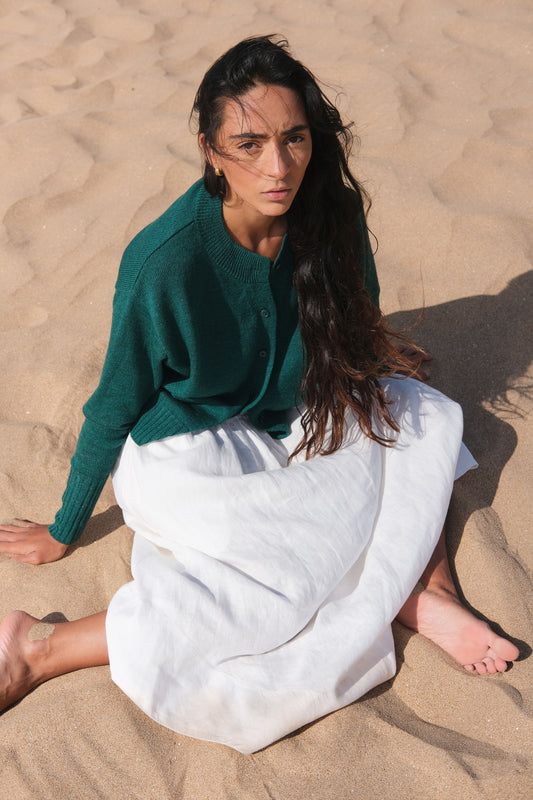 LILLY PILLY woman wearing pine green coloured Taylor merino knit cardigan sitting on sand wearing a green sweater and white skirt