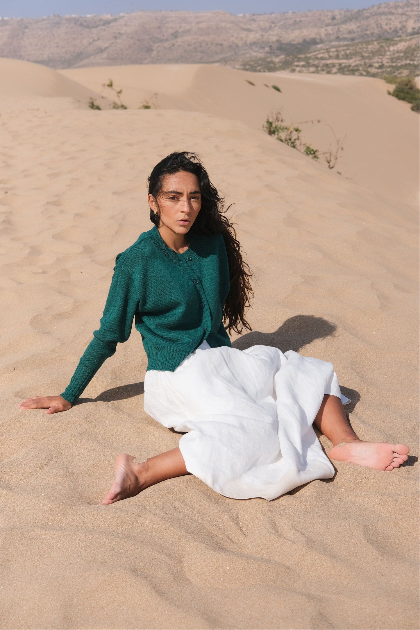 LILLY PILLY woman wearing pine green coloured Taylor merino knit cardigan  sitting on sand dunes wearing a green sweater and white skirt.