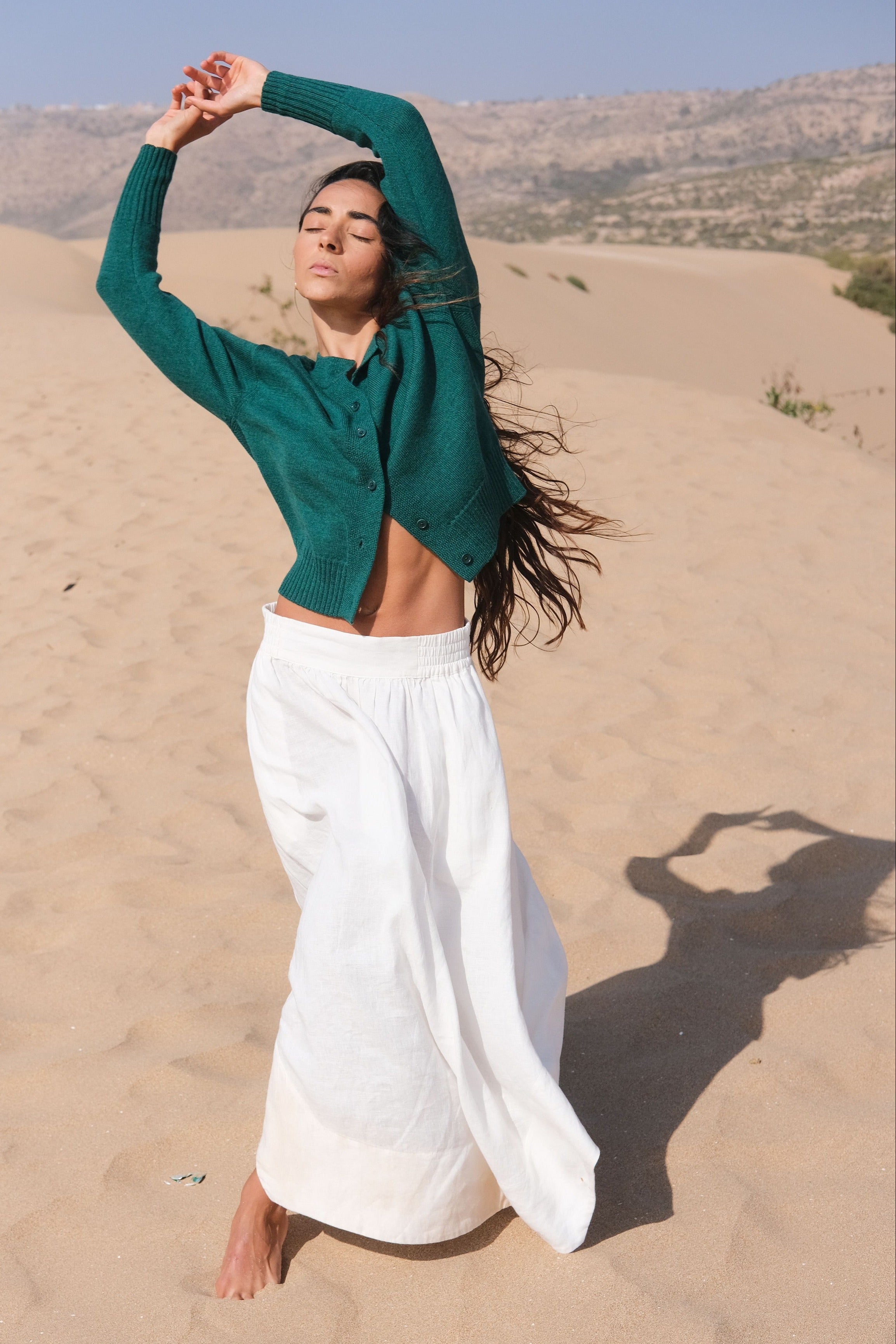 LILLY PILLY woman wearing pine green coloured Taylor merino knit cardigan  standing in a desert landscape with mountains in the background.