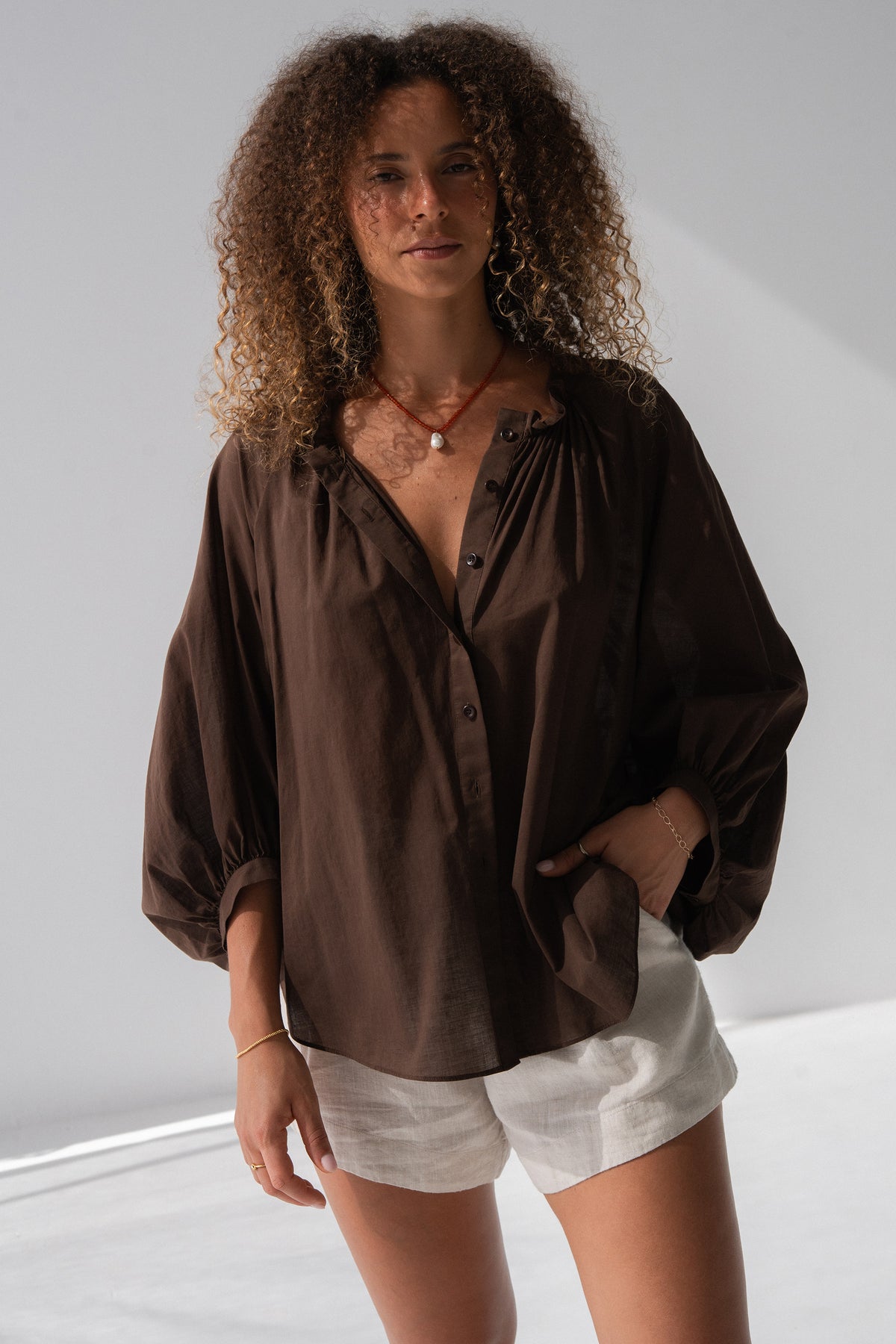 Thalia Cotton Voile Shirt in Chocolate - front view, gathered neckline and button-front, balloon sleeves, hand in pocket, worn over Gaia Linen Shorts in Oatmeal - Lilly Pilly Collection