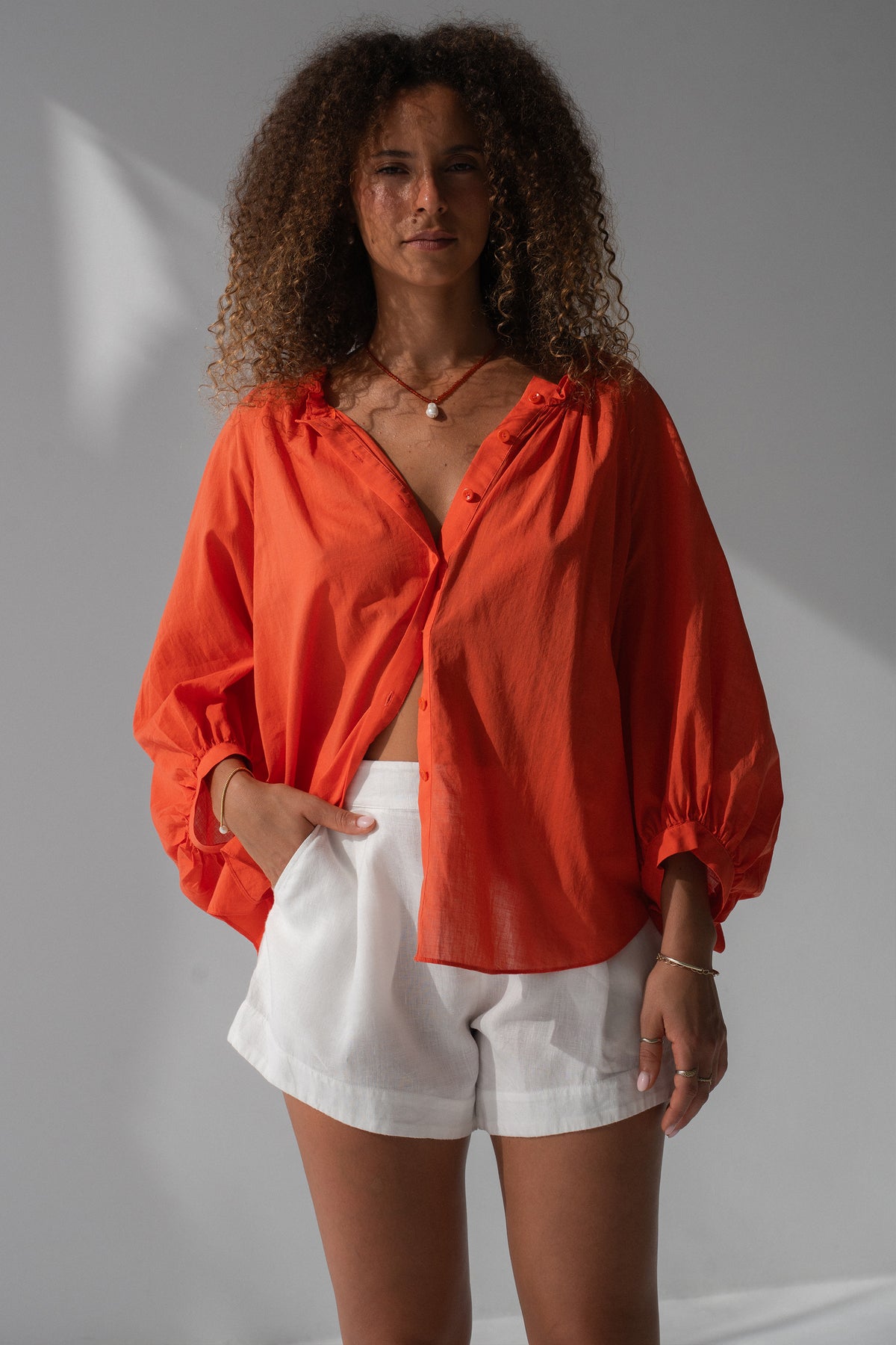 Thalia Cotton Voile Shirt in Spritz Orange - full length front view, gathered neckline and button-front, balloon sleeves, worn open over white linen shorts, hand in pocket - Lilly Pilly Collection