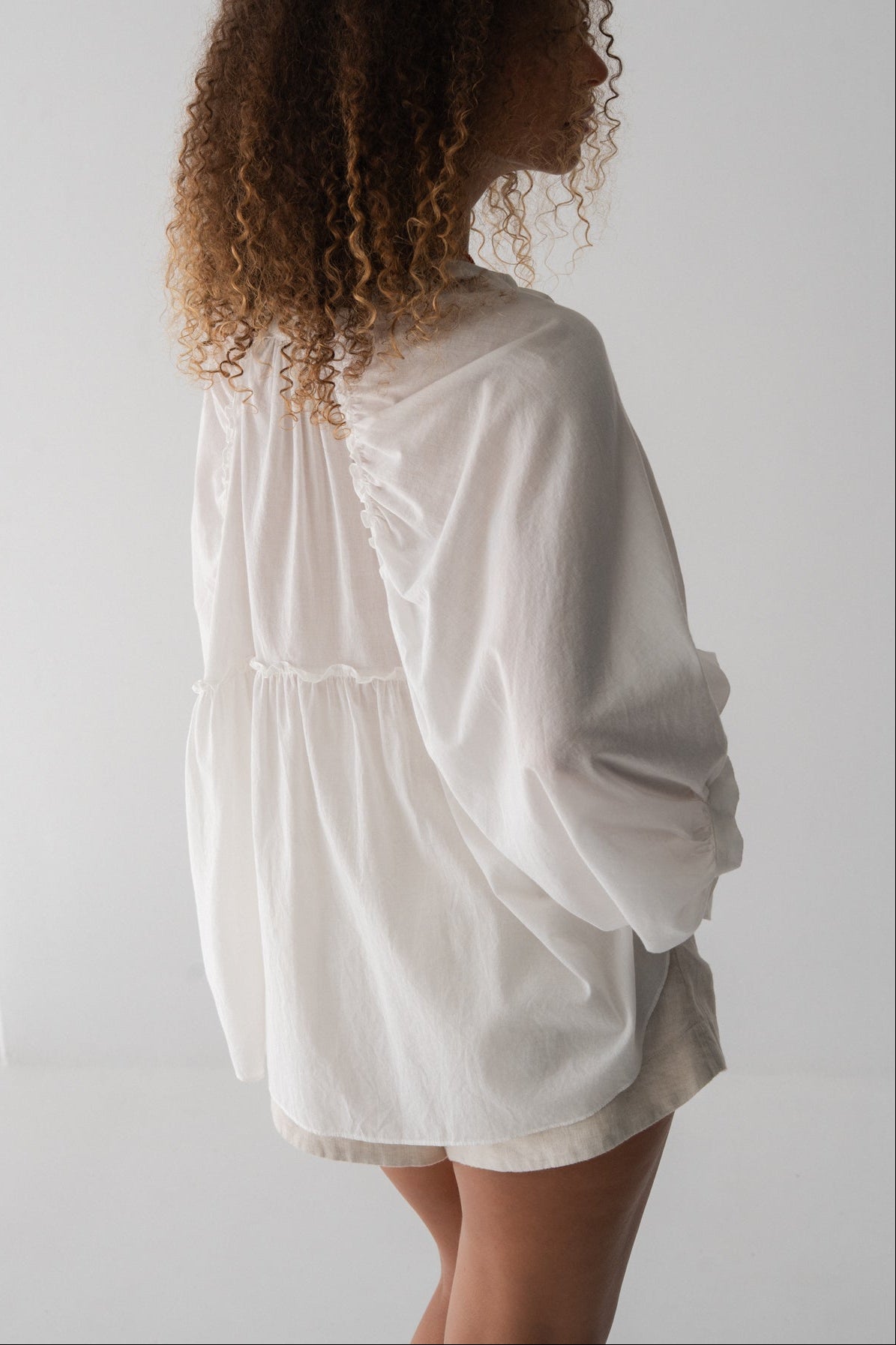 Close up side view of LILLY PILLY woman wearing a ivory Thalia cotton voile shirt blouse and oatmeal-coloured Gaia shorts against a neutral background