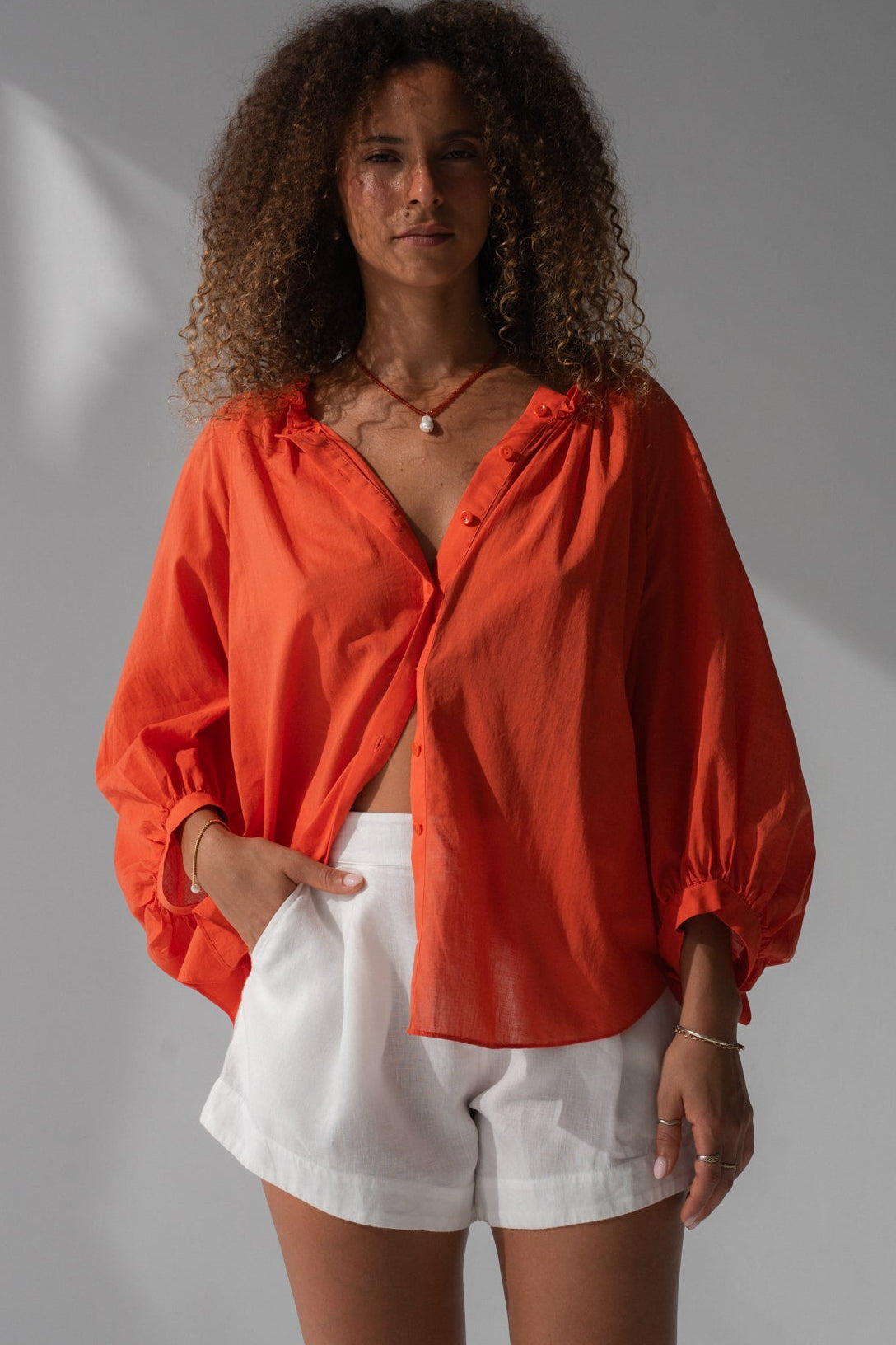 LILLY PILLY person wearing an unbuttoned spritz cotton voile Thalia top and ivory white Gaia linen shorts against a grey background