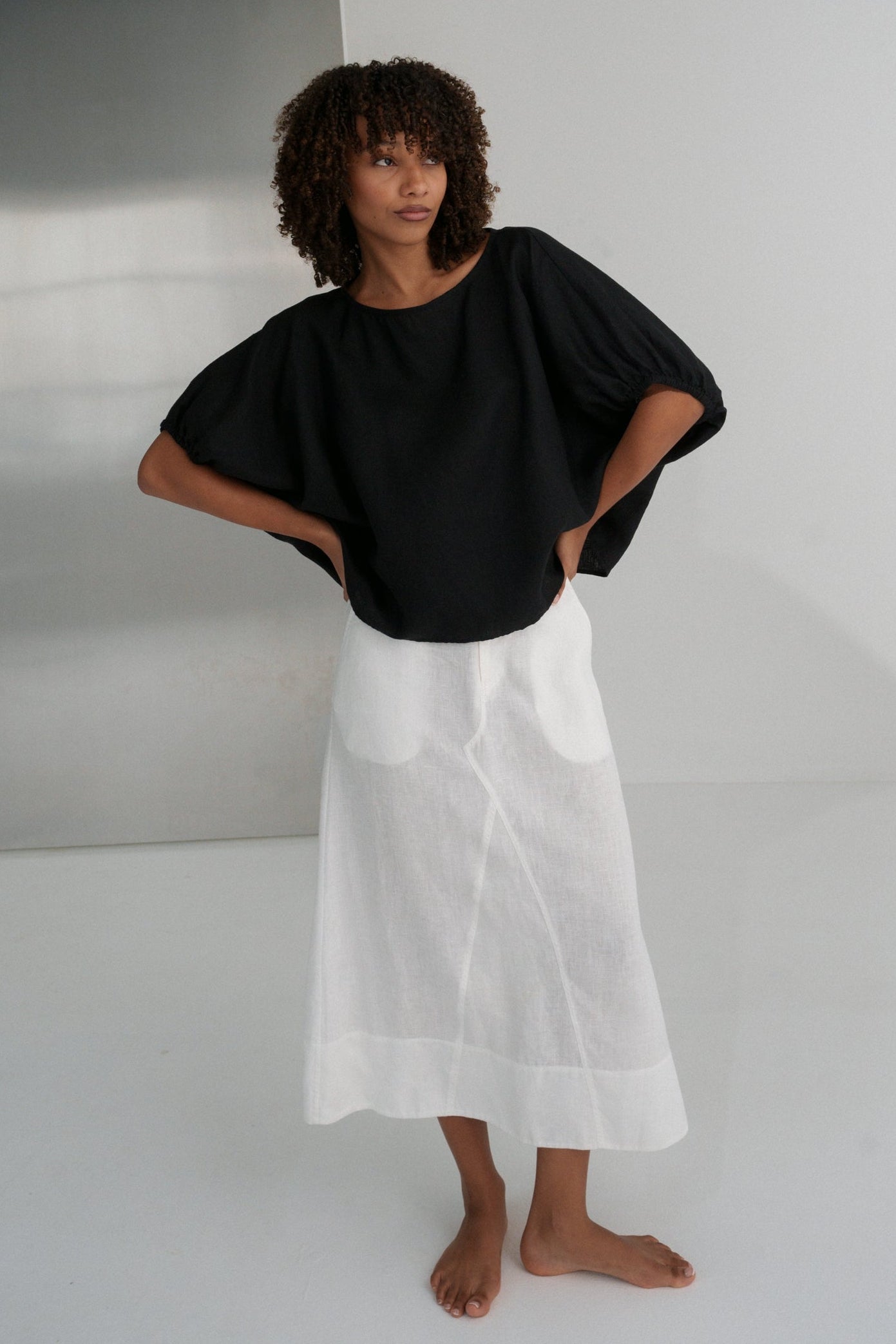 LILLY PILLY woman in an black Tina linen top and ivory linen skirt standing against a white wall.