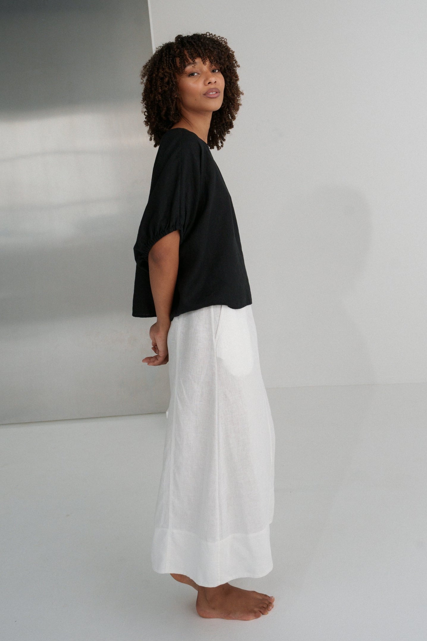 Side view of LILLY PILLY woman in an black Tina linen top and ivory linen skirt standing against a plain white wall.