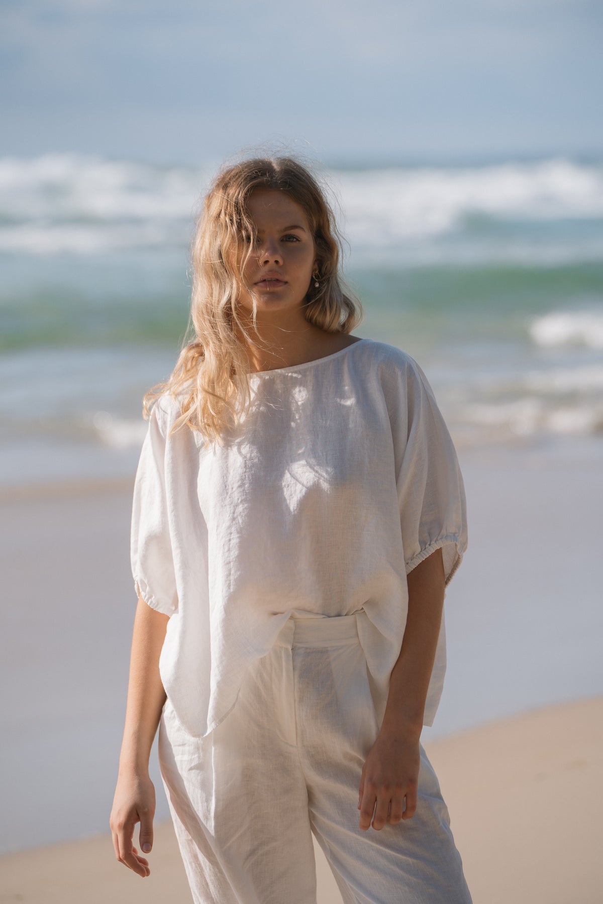 Tina Linen Top in Ivory - front view, boat neckline and elasticated cuffs, standing on the beach - Lilly Pilly Collection