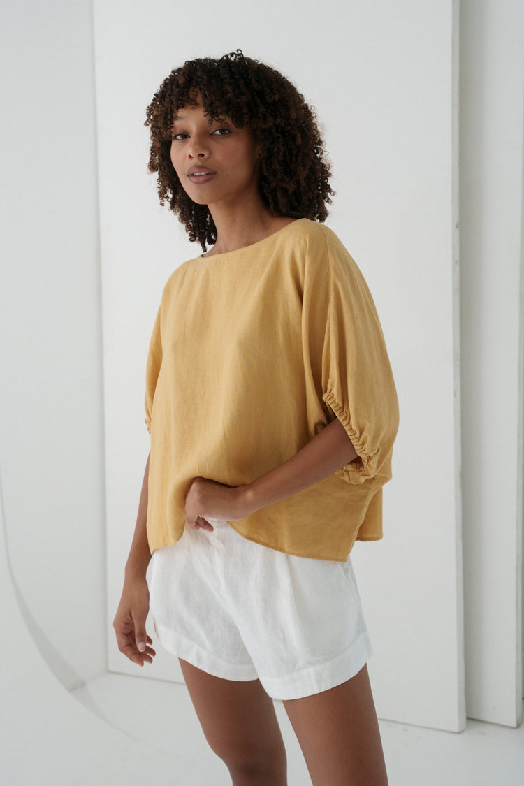 Tina Linen Top in Sand - three-quarter view, round neckline and batwing sleeves with elasticated cuffs, styled with white linen shorts - Lilly Pilly Collection