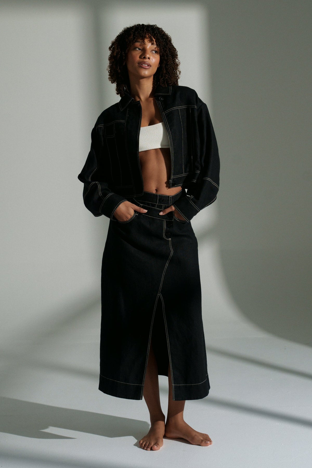 LILLY PILLY woman wearing a black Wes linen jacket standing in a minimalistic setting with light casting shadows.