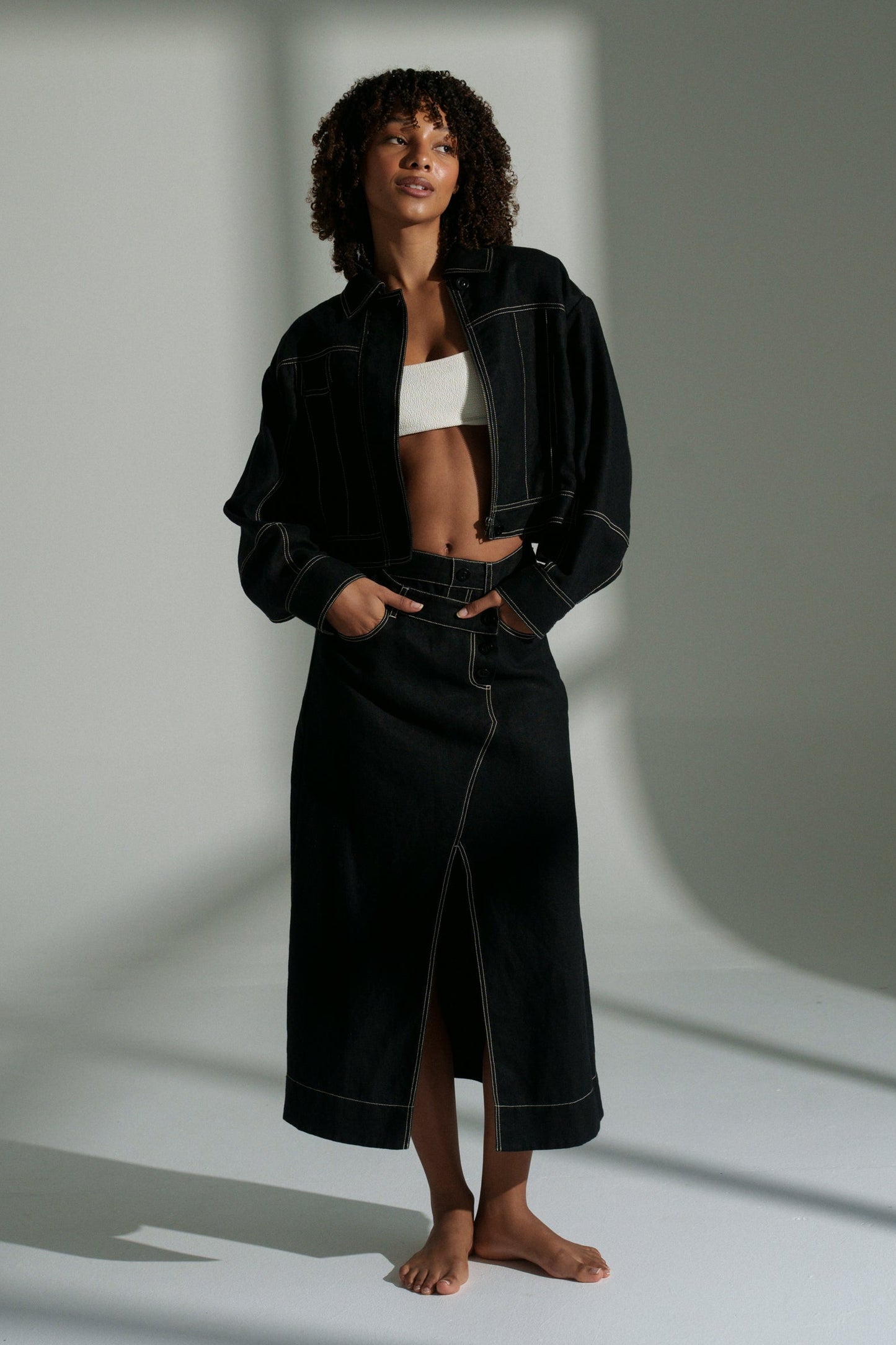 LILLY PILLY woman wearing a black Wes linen jacket standing in a minimalistic setting with light casting shadows.