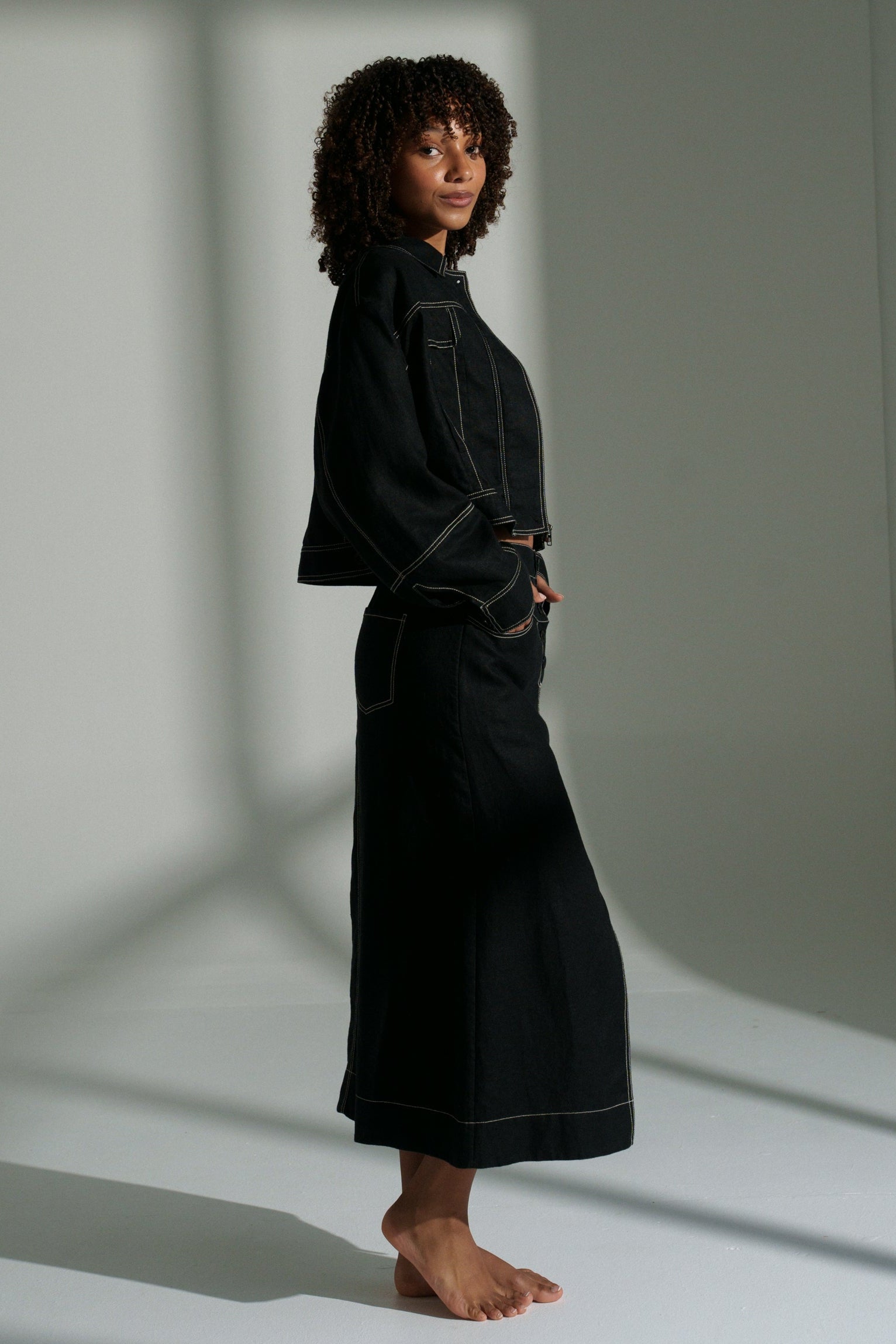 LILLY PILLY woman wearing a black Wes linen jacket standing in a room with light casting shadows on the wall.