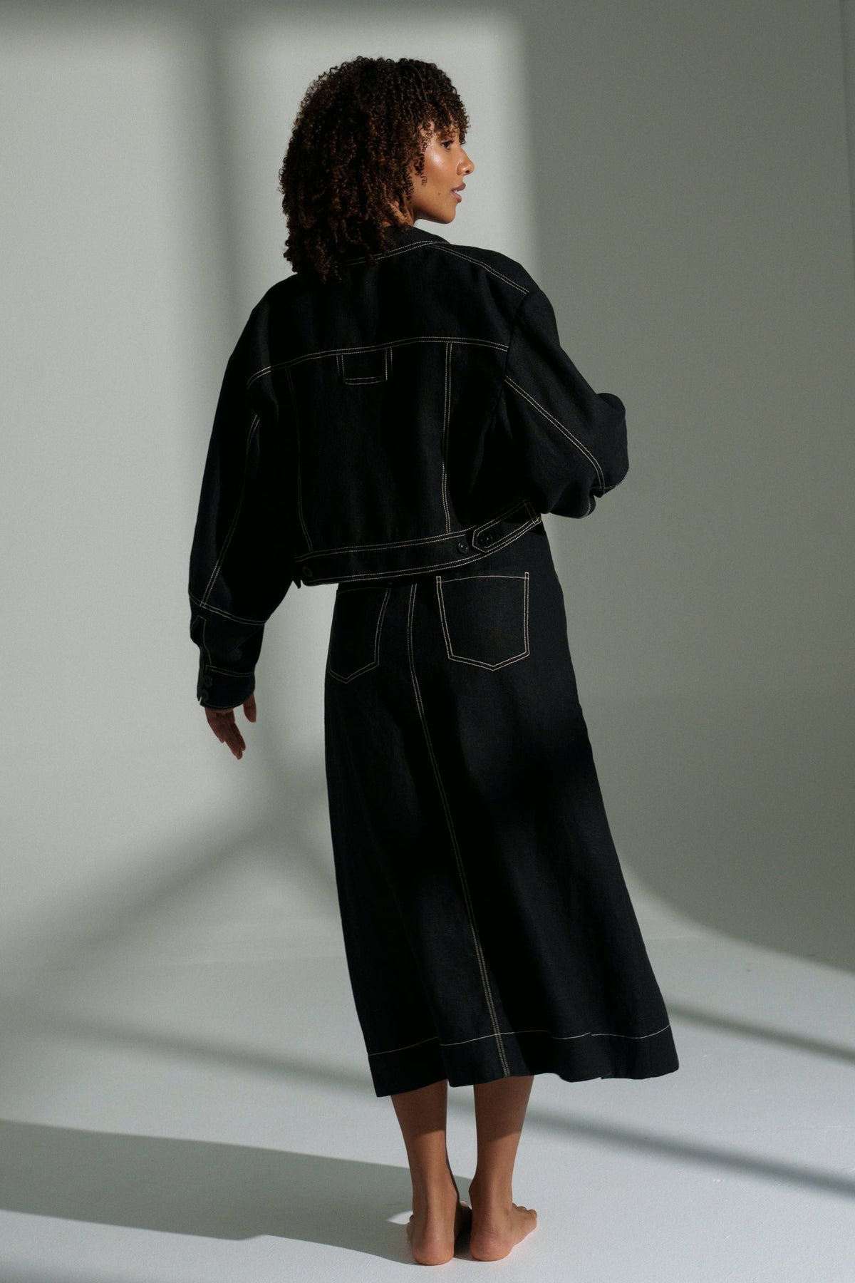 LILLY PILLY woman wearing a black Wes linen jacket and Jax skirt outfit standing in a minimalistic room with light casting shadows.