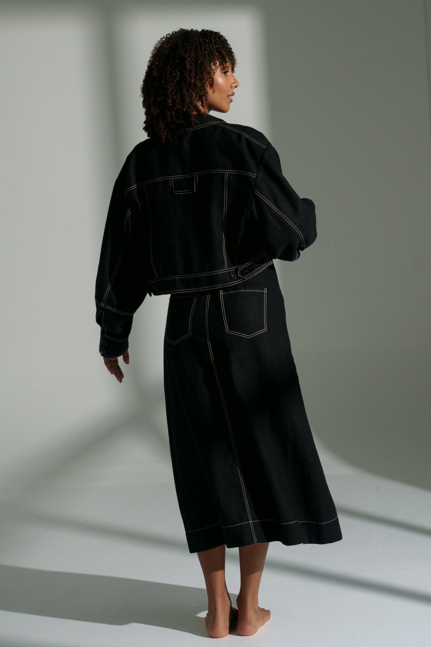 LILLY PILLY woman wearing a black Wes linen jacket and Jax skirt outfit standing in a minimalistic room with light casting shadows.