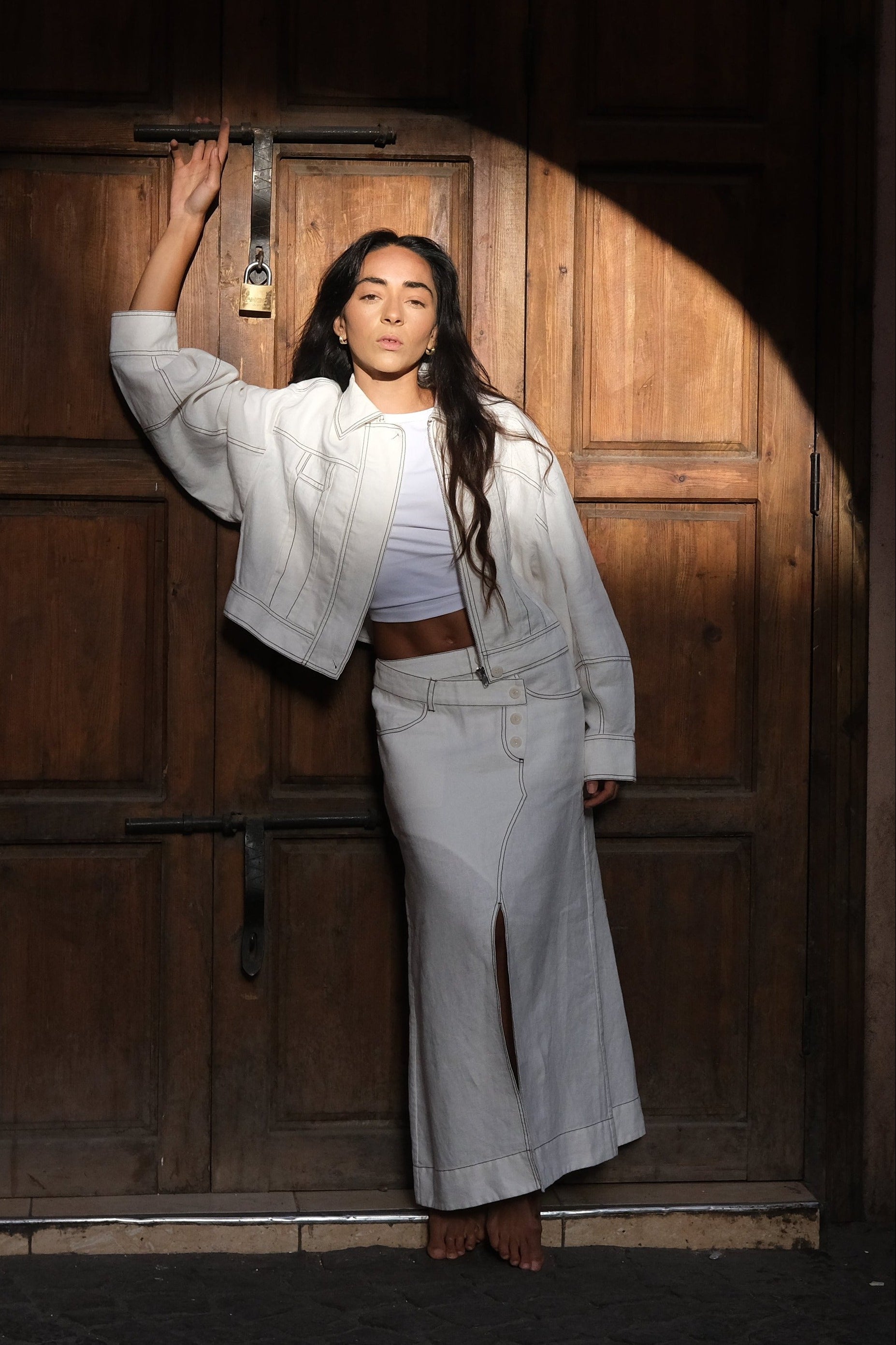 LILLY PILLY woman wearing an ivory Wes linen jacket standing in front of a wooden door with warm lighting.