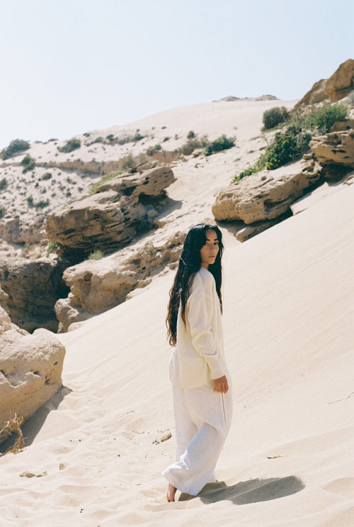 LILLY PILLY woman in an ivory June cashmere knitwear sweater and white linen pants standing in a desert landscape with rocky formations.
