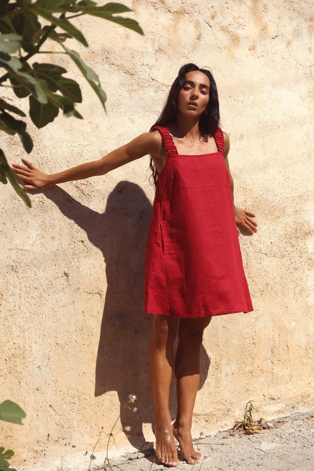 Zahra Linen Mini Dress in Cherry Red - three-quarter view against sandstone wall, ruched straps and relaxed A-line silhouette - Lilly Pilly Collection