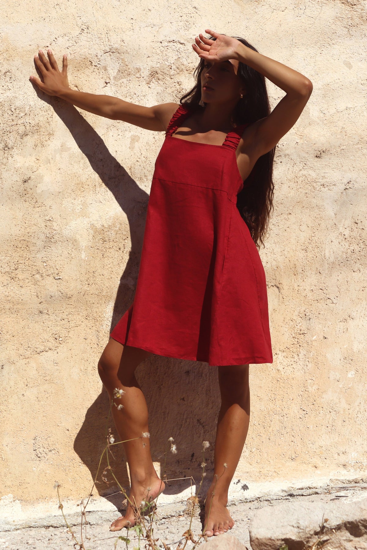 Zahra Linen Mini Dress in Cherry Red - woman against sandstone wall, arms raised, ruched straps and A-line skirt - Lilly Pilly Collection