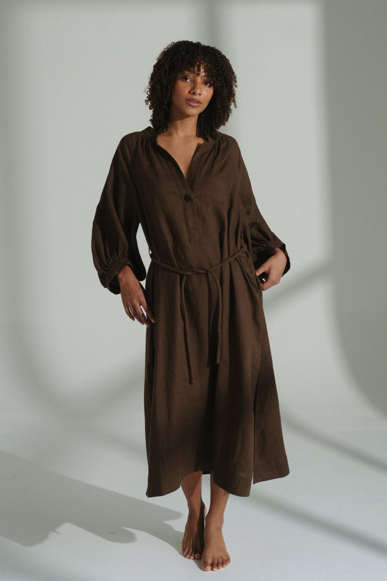 LILLY PILLY woman wearing a chocolate brown Mae linen dress standing in a softly lit room with shadows on the wall.