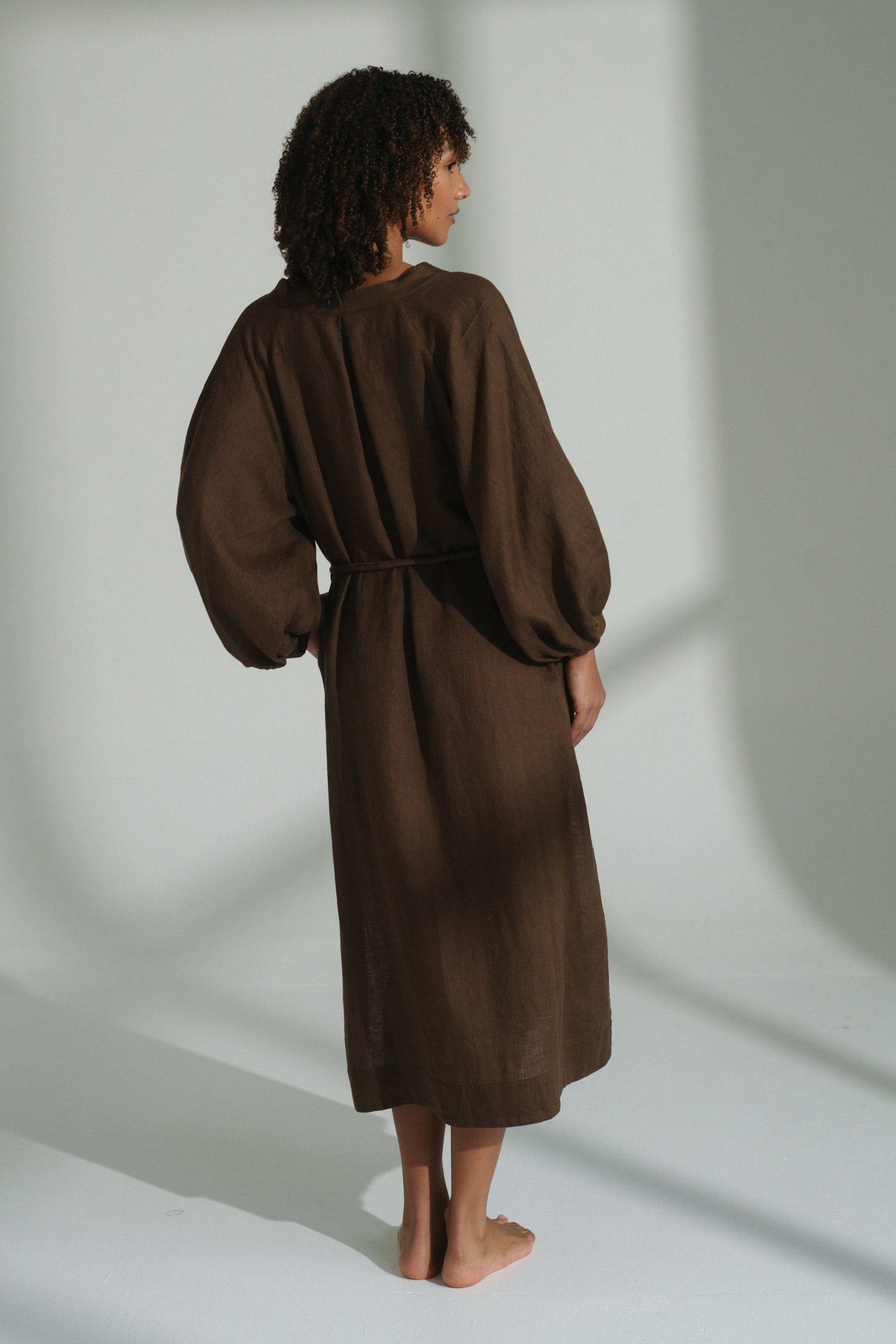 LILLY PILLY woman  wearing a chocolate brown Mae linen dress standing in a softly lit room with shadows on the wall.
