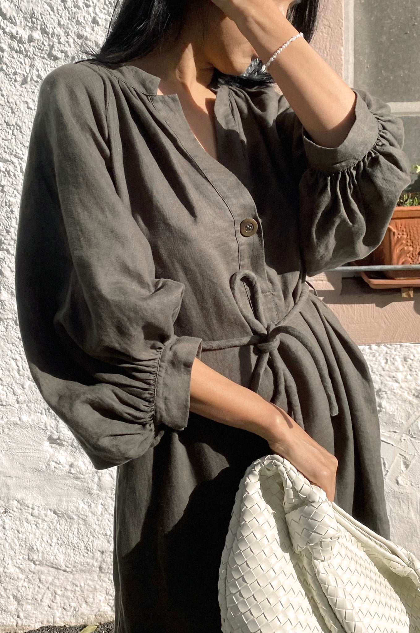 LILLY PILLY woman wearing a Mae khaki linen dress with a white textured bag, standing against a light-colored wall.