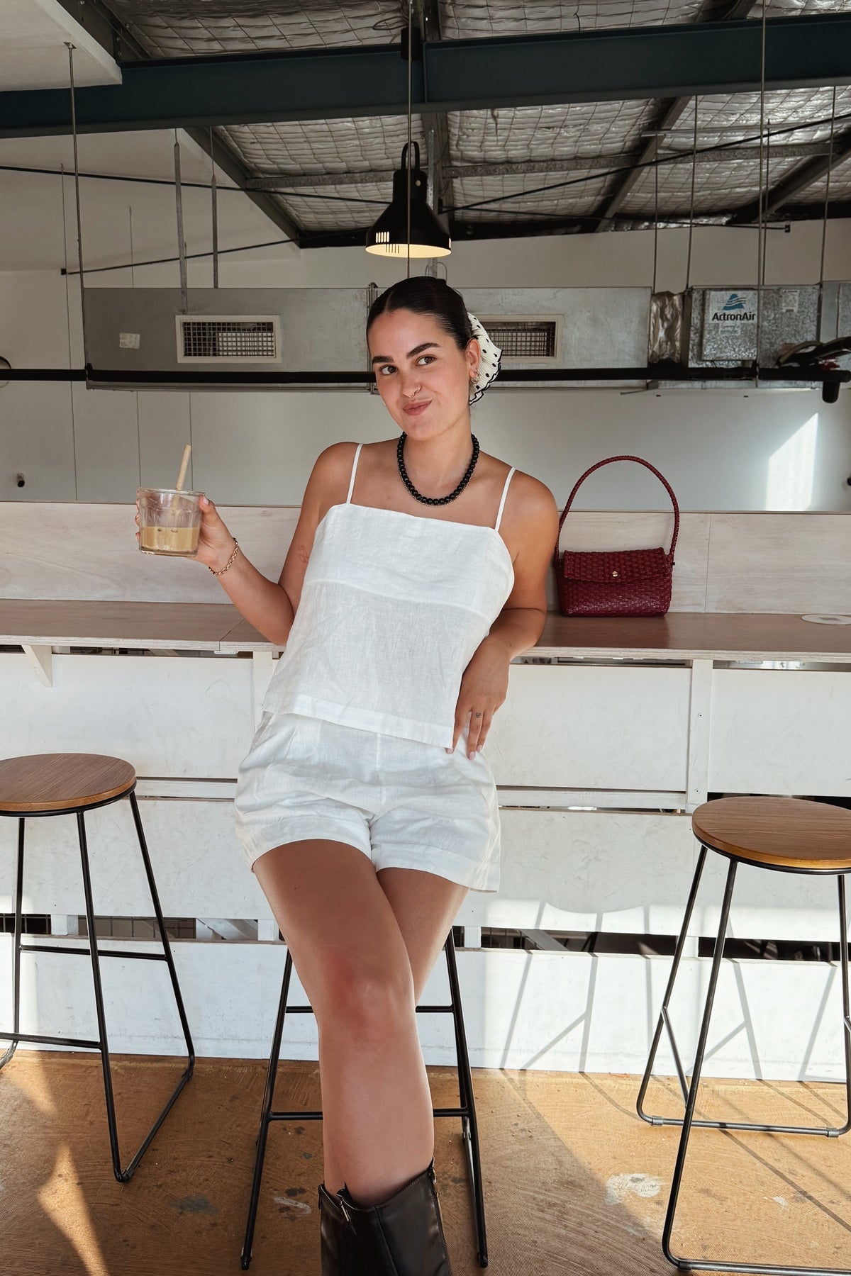 LILLY PILLY woman in ivory Rosa linen cami and linen Gaia shorts sitting at a bar holding a drink.