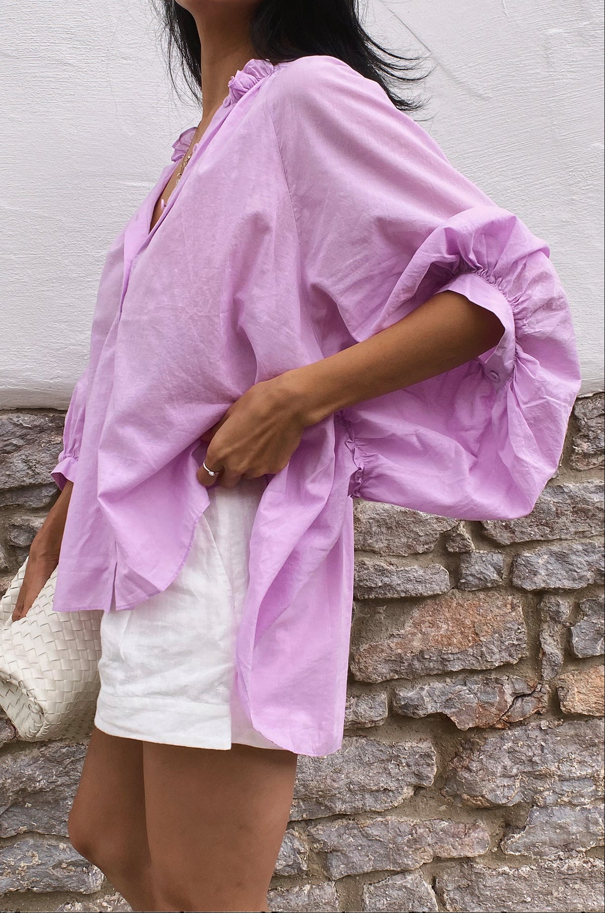 Close up side view of LILLY PILLY woman wearing a orchid Thalia cotton voile shirt  while walking past a textured rock wall.