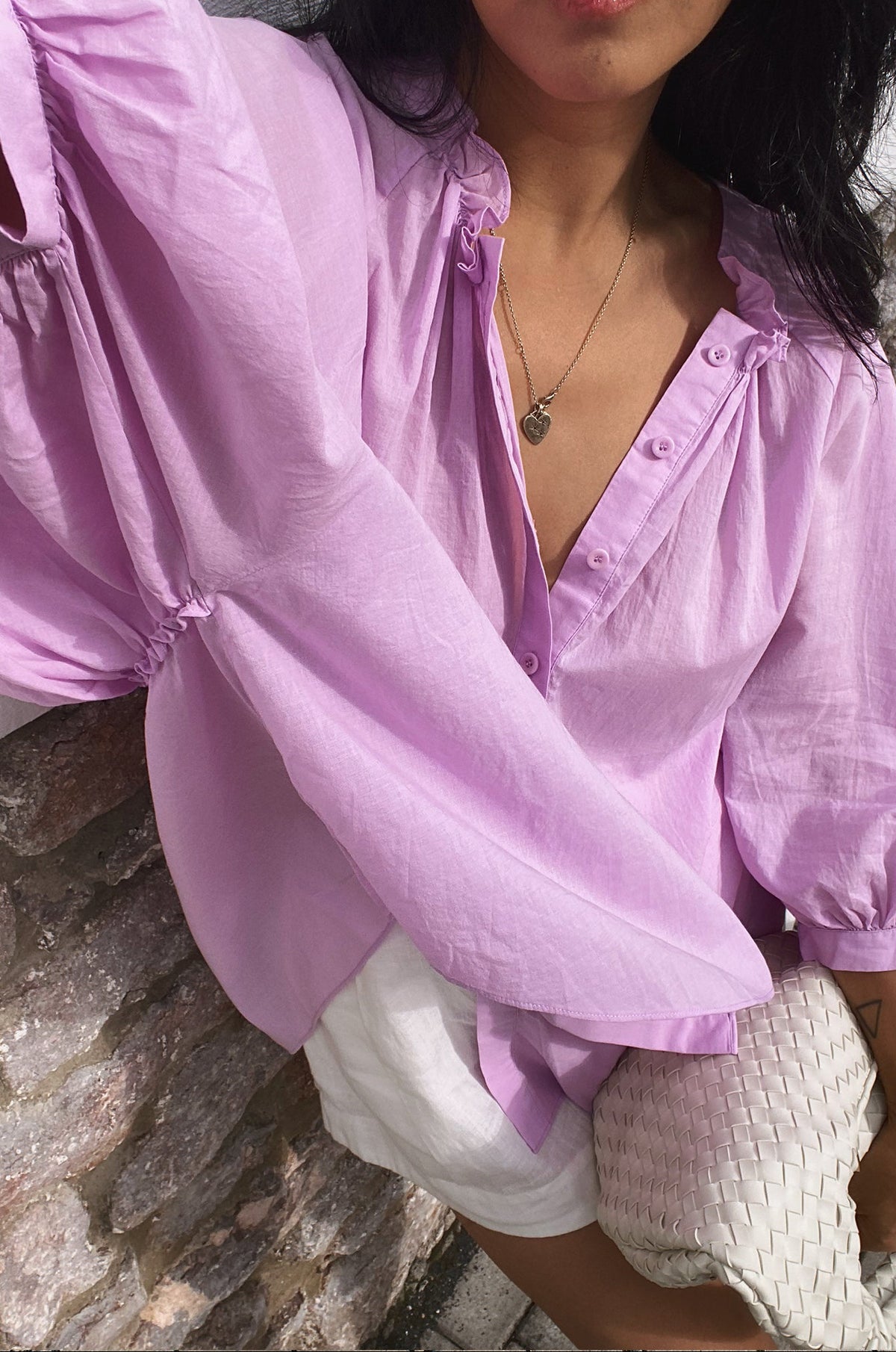 Close up front view of collar details of woman wearing a orchid Thalia cotton voile shirt standing by a textured rock wall.