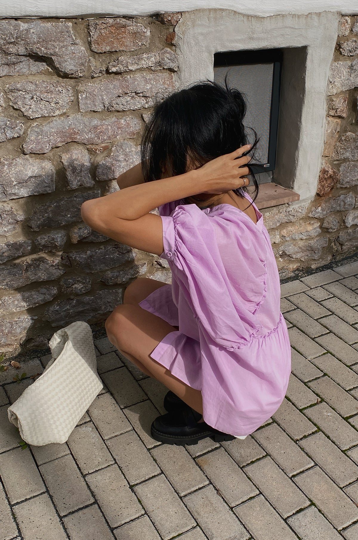 Close up side view of woman wearing a orchid Thalia cotton voile shirt crouching down next to a stone wall.