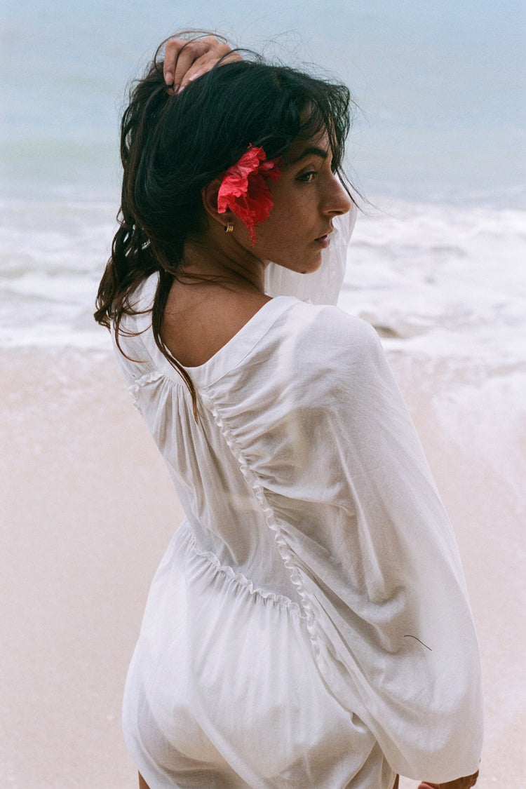 Thalia Cotton Voile Shirt in Ivory - back view, gathered back yoke and balloon sleeves, hibiscus flower, worn as beach cover-up, ocean backdrop - Lilly Pilly Collection